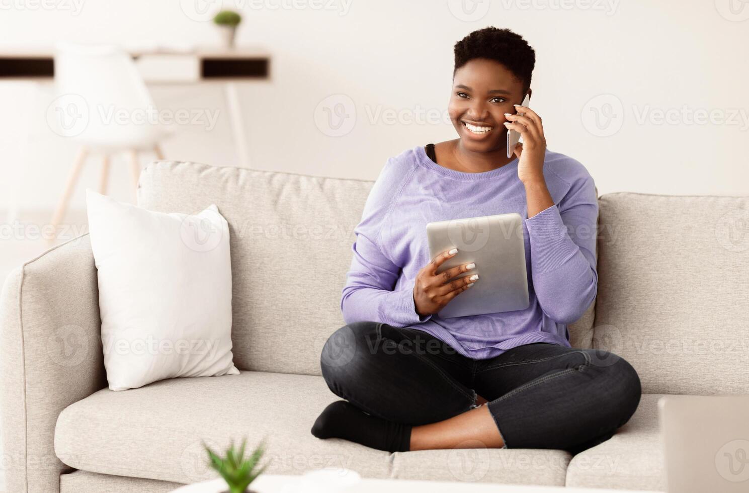 Always In Touch. Happy young plus size african american woman talking on mobile phone, sitting on the couch at home and holding digital tablet. Communication And Technology Concept. photo