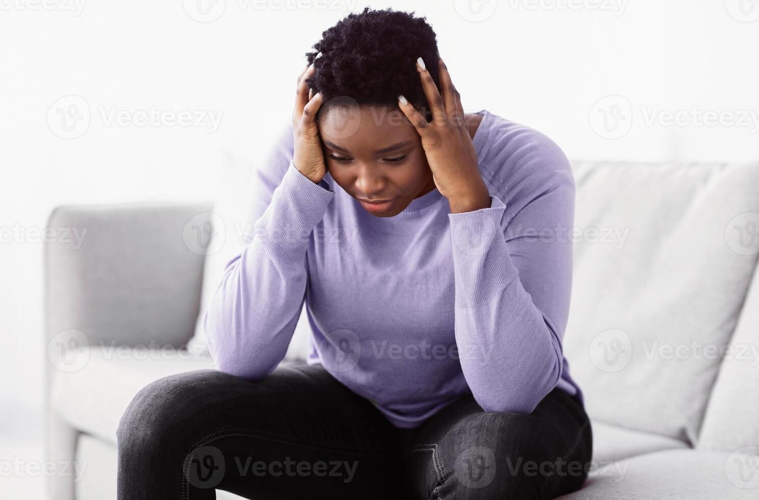 Portrait of exhausted black young woman grabbing her head feeling stress, suffering from pain or pressure, having problems, thinking about troubles at work, sitting on the couch at home photo