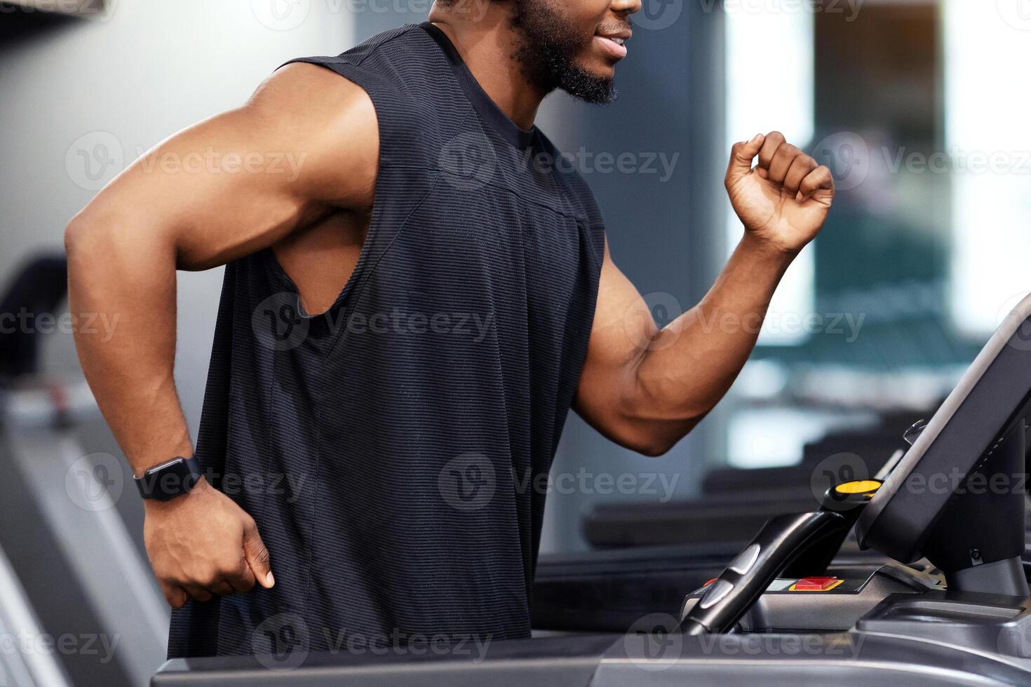Jogging workout. Joyful black man in modern wireless headphones running on treadmill at gym, looking at free space. Motivated african american guy in black sportswear spending time at gym, jogging photo