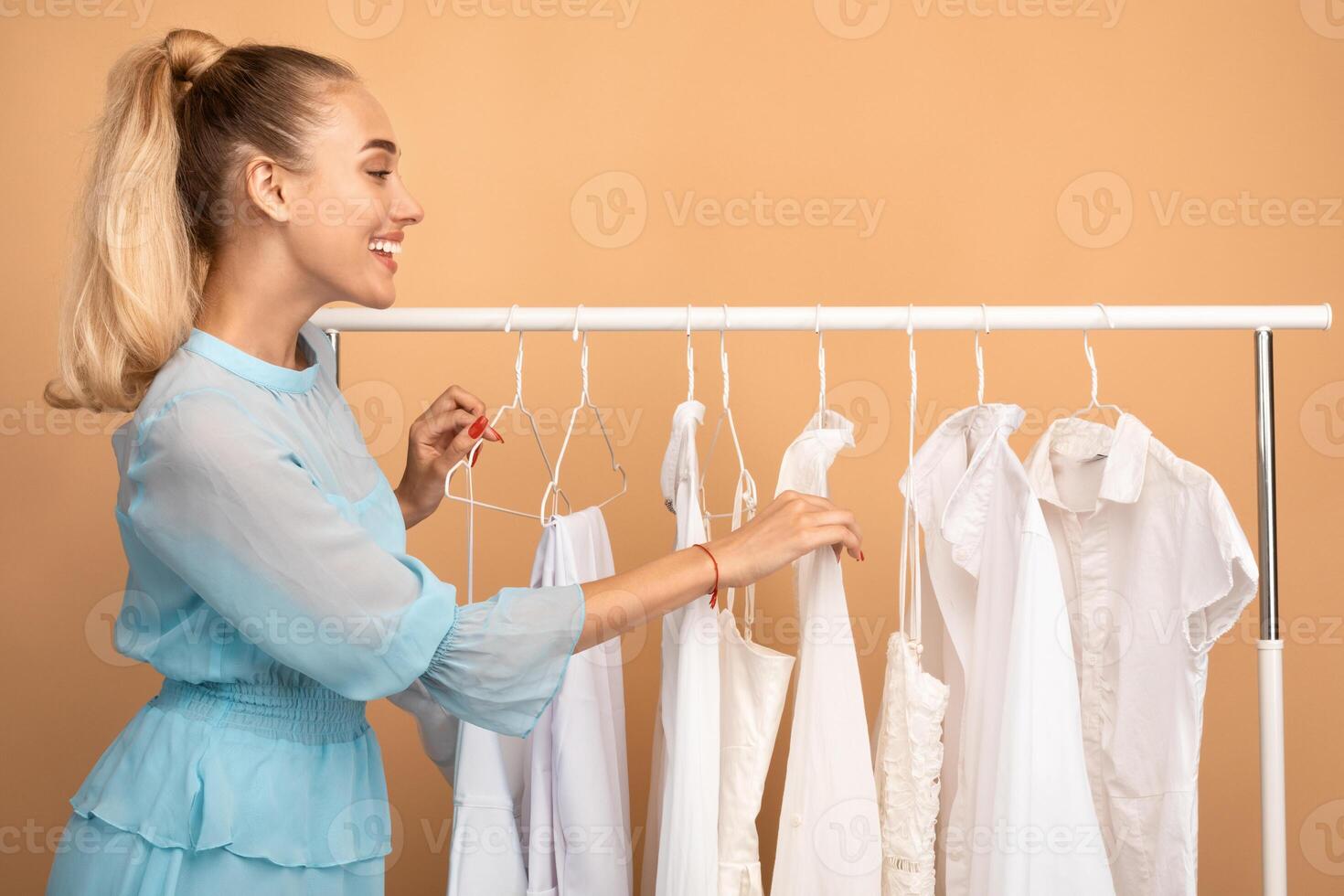 New Collection. Cheerful young woman shopping in fashion mall, choosing new clothes, customer looking through hangers with stylish casual garment on clothing rail, beige studio background photo