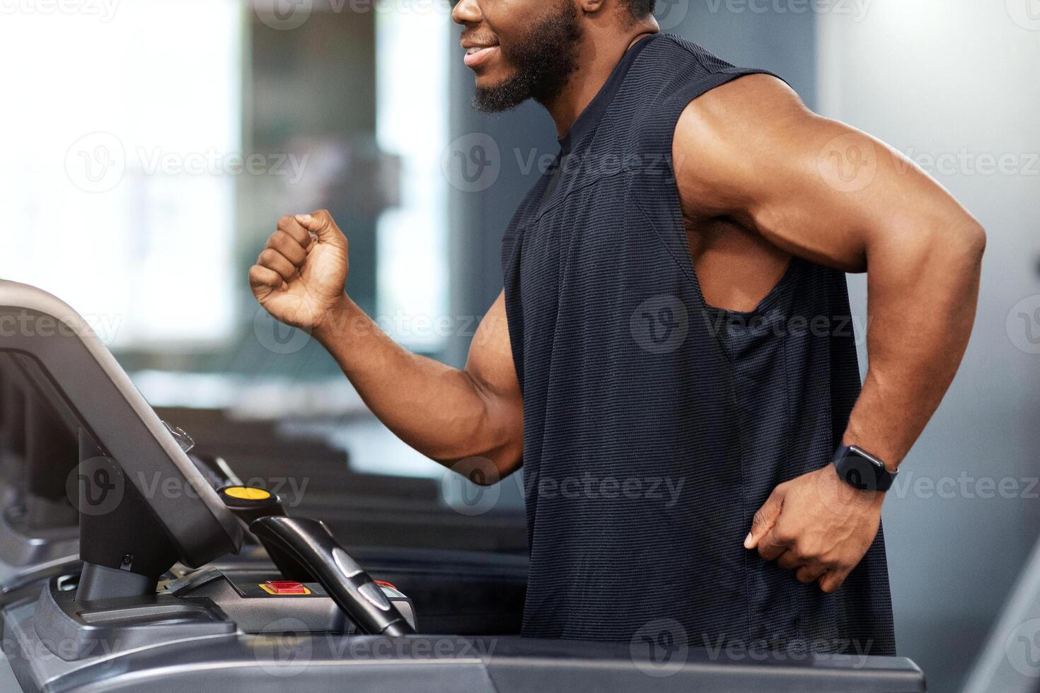 Motivated african american guy in black sportswear spending time at gym, jogging, warming up before work out. Side view of happy black man running on treadmill, looking at free space photo