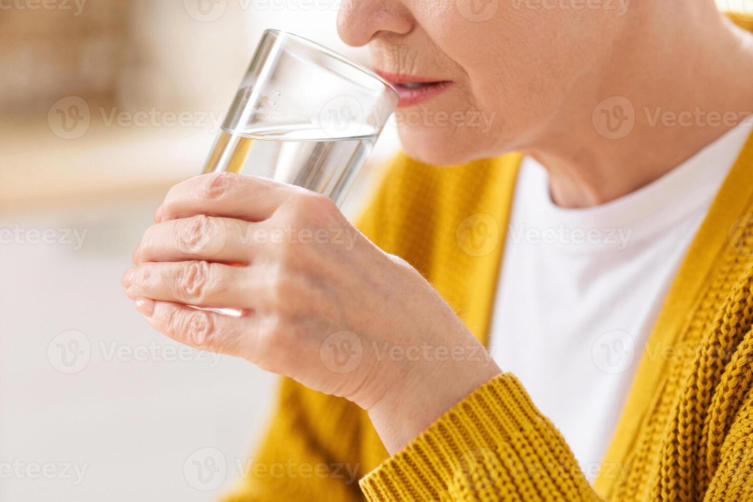 Health benefits of drinking water concept. Closeup of glass with spring water in old woman hand, selective focus. Unrecognizable grandmother drinking still fresh water, cropped, empty space photo
