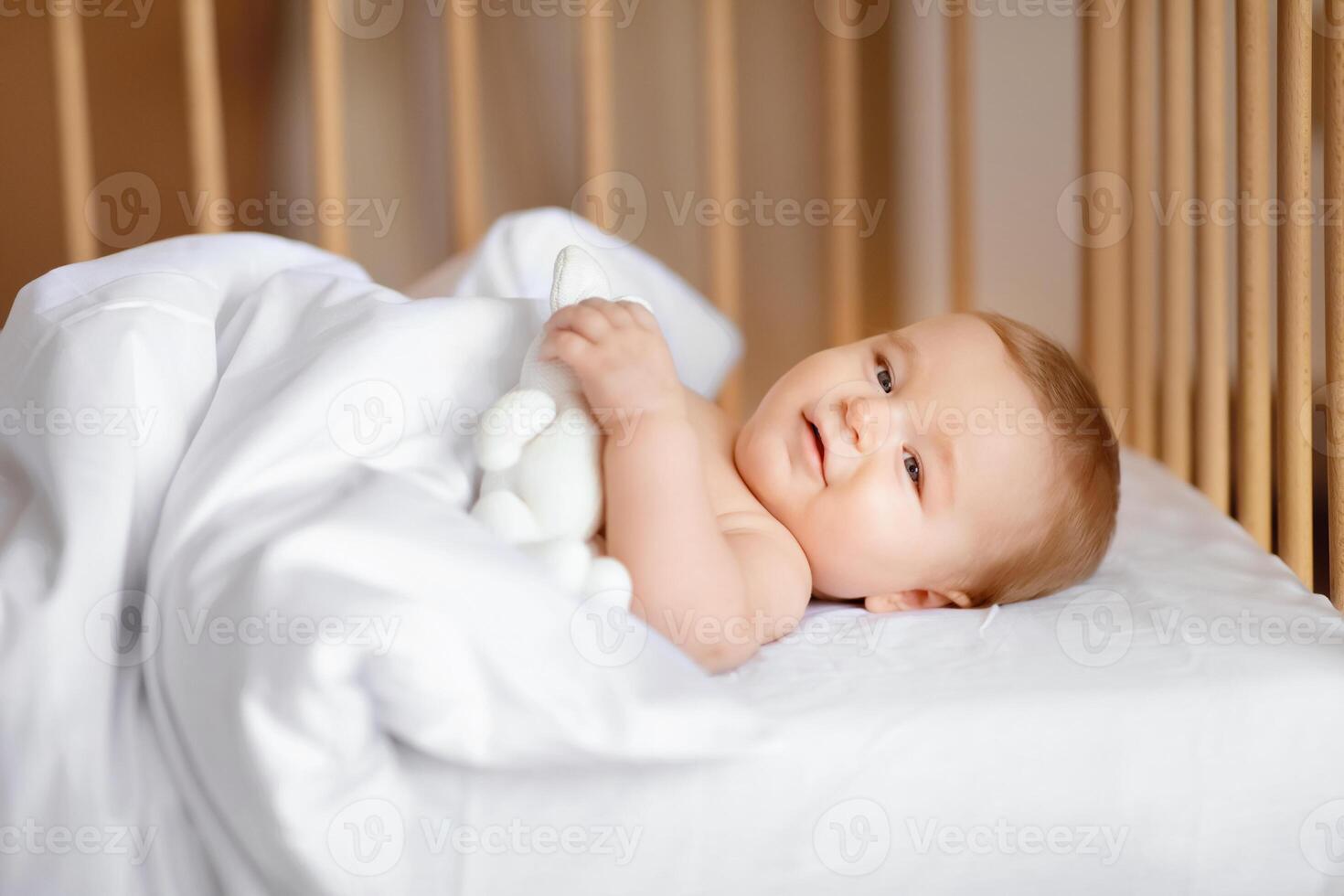 Good morning world. Happy baby boy waking up after sleeping in wooden crib, laying under blanket and smiling, holding white toy, side view. Cute baby girl laying in toddler bed photo