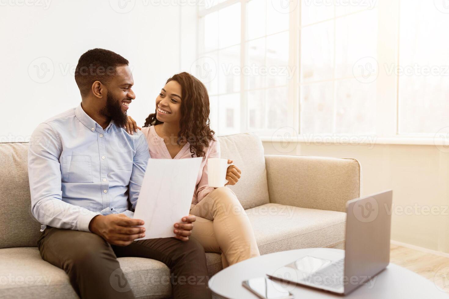 Planning Family Budget. Smiling african american man holding paper, working and reading documentation at home, woman drinking coffee and looking at him, sitting on the couch with laptop photo