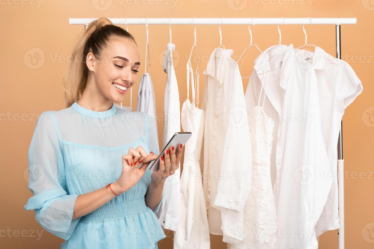 Sales And Discounts Season. Portrait of smiling young woman holding and using her mobile phone, chatting or making purchases in online store standing near clothing rail at the studio photo