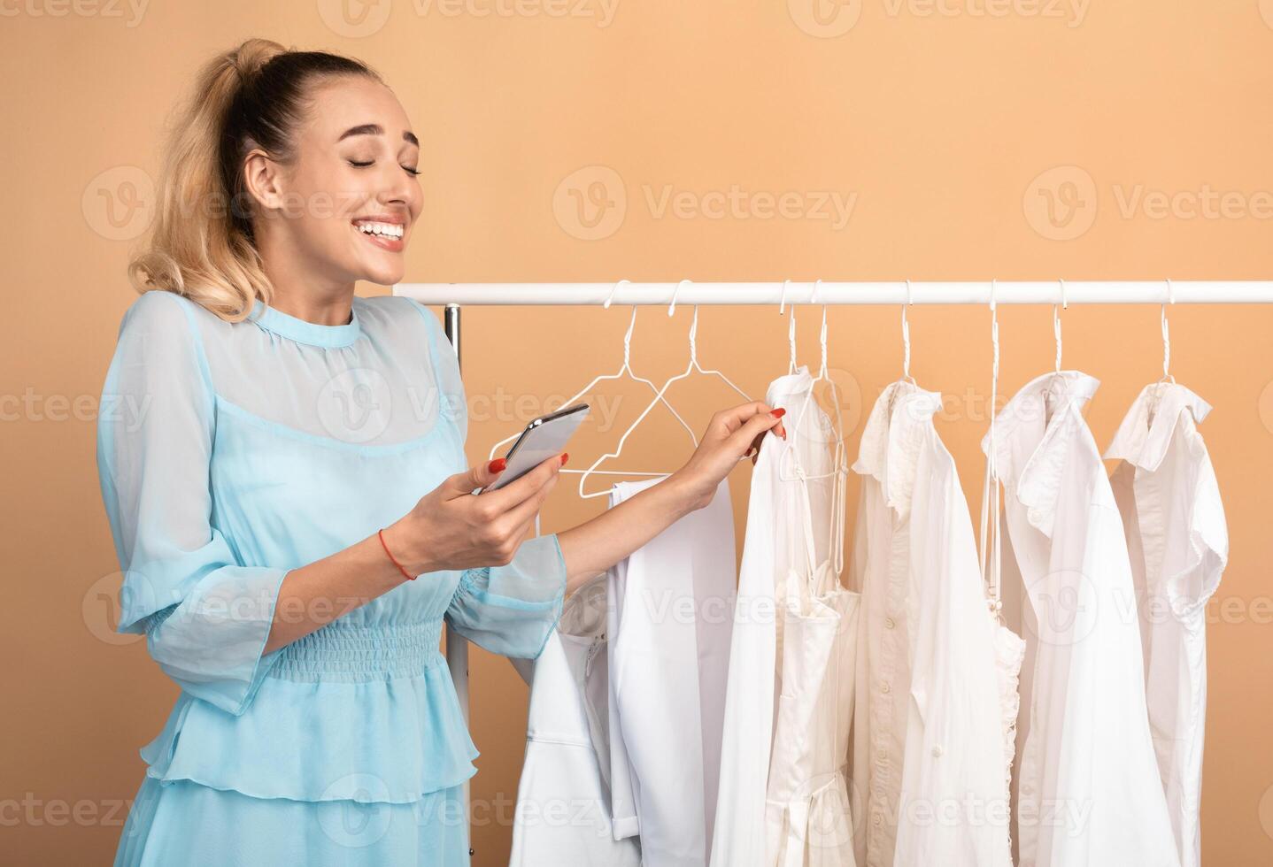 Portrait of emotional female customer holding and using mobile phone, feeling excited, texting sms or making purchases in online store standing near rack with clothes at the mall photo