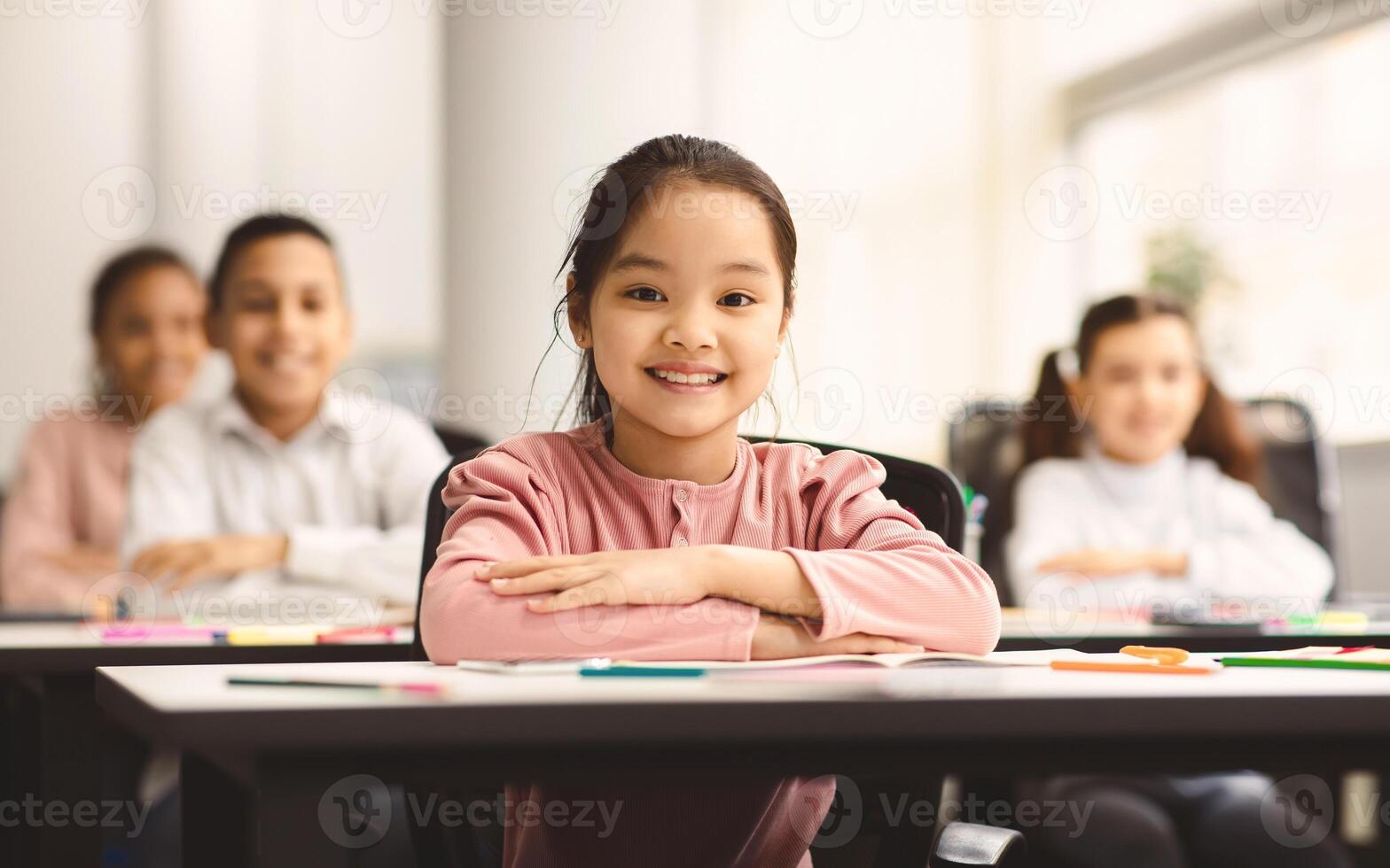 First Day At School. Portrait of smiling diligent asian girl sitting with folded arms at table with group of classmates in blurred background, posing, looking at camera, selective focus. Back To Study photo