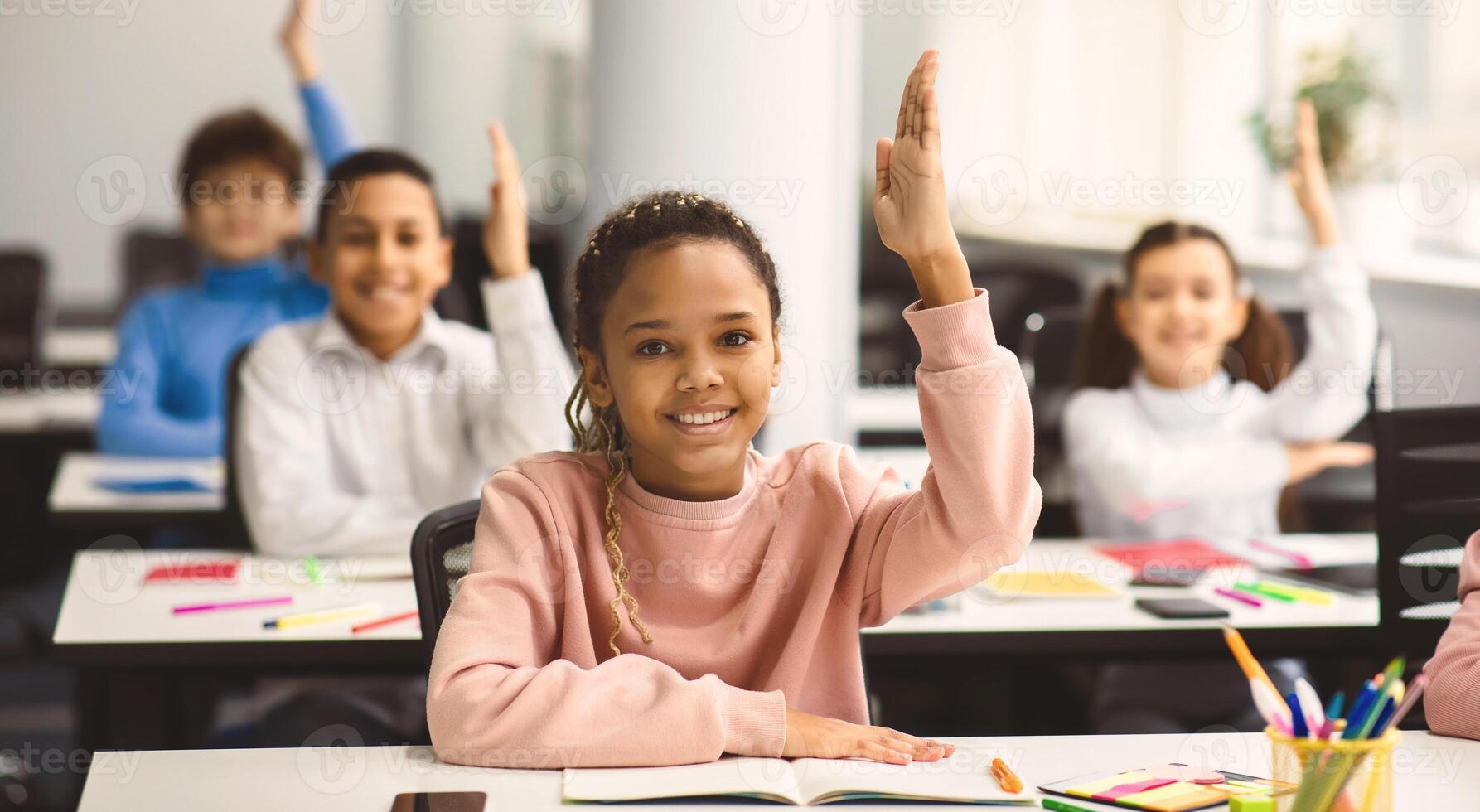 Education, Primary School, Learning and People Concept. Happy black schoolgirl raising hand for an answer, sitting at desk in classroom with diverse group of smiling classmates, studying with pleasure photo