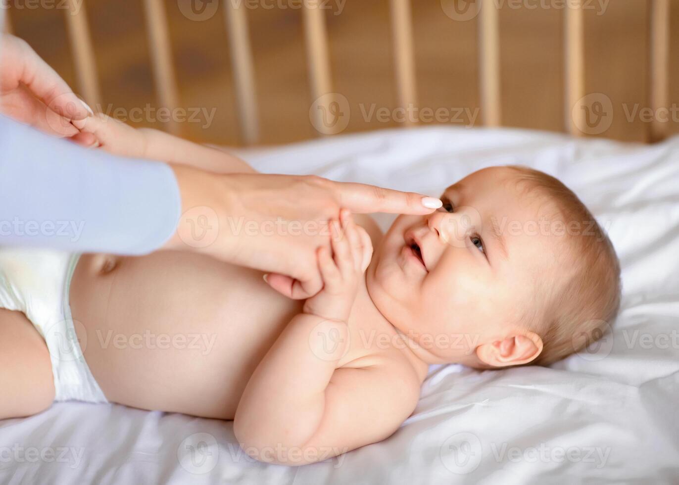 Closeup of adorable toddler playing with mom after waking up, laying in wooden baby bed and smiling. Unrecognizable mother touching her cute happy laughing baby boy or girl nose photo