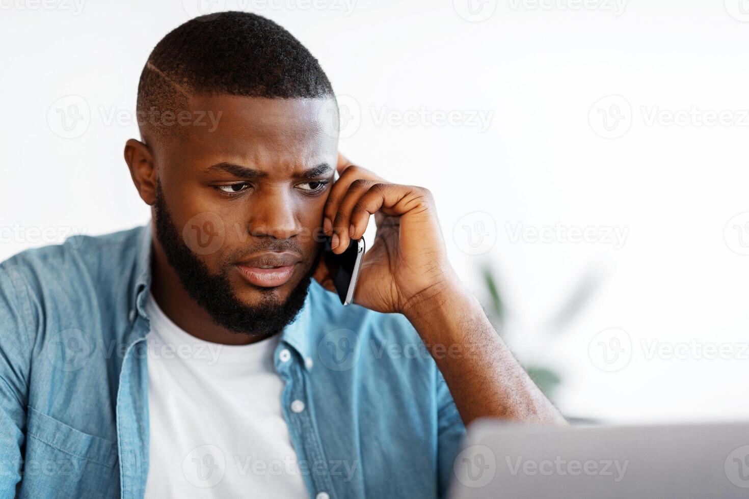 Portrait Of Busy African American Man Talking On Cellphone And Working On Laptop, Millennial Black Male Entrepreneur Looking At Computer Screen And Having Phone Conversation, Closeup photo
