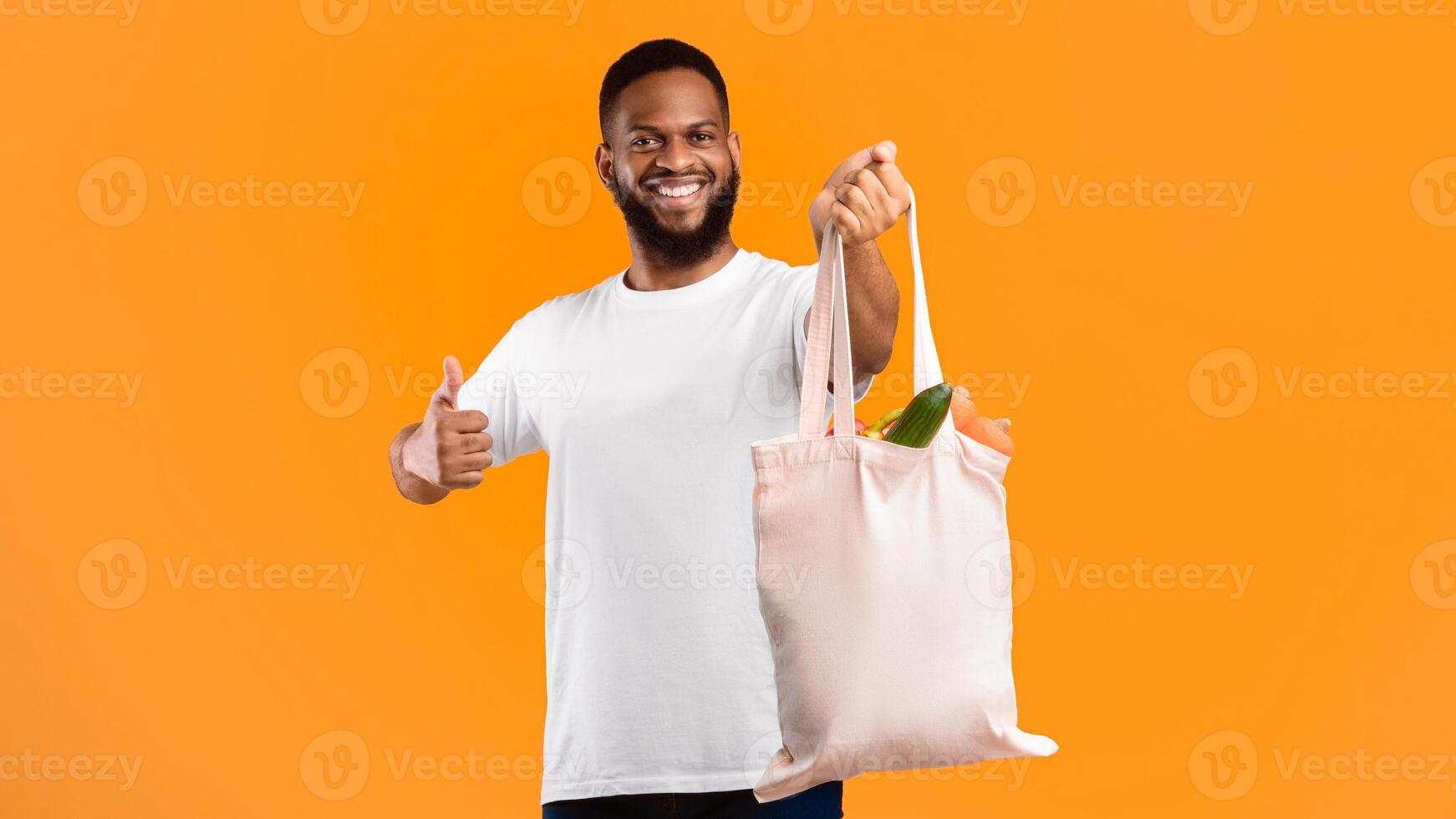 Plastic-Free Life. African Man Holding Eco Bag With Groceries And Gesturing Like Smiling To Camera Standing Over Yellow Studio Background. I Like Eco-Friendly Lifestyle, Ecology And Consumerism photo