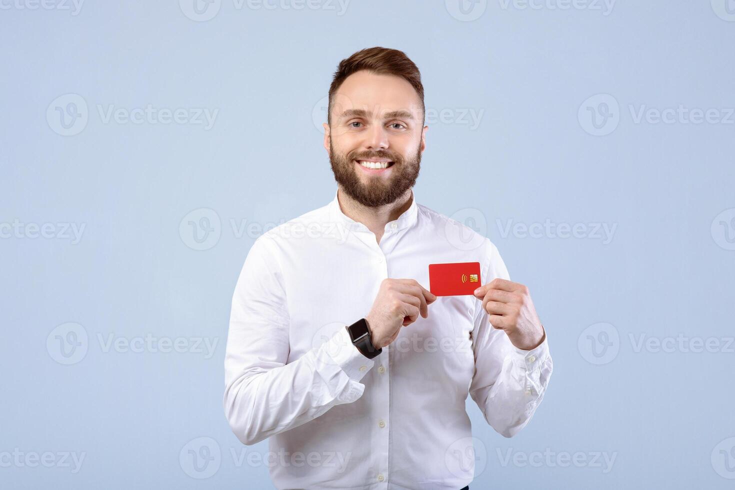 Cool young man in office wear holding credit card and smiling at camera on blue studio background. Satisfied bank client promoting financial product, choosing contactless payment photo