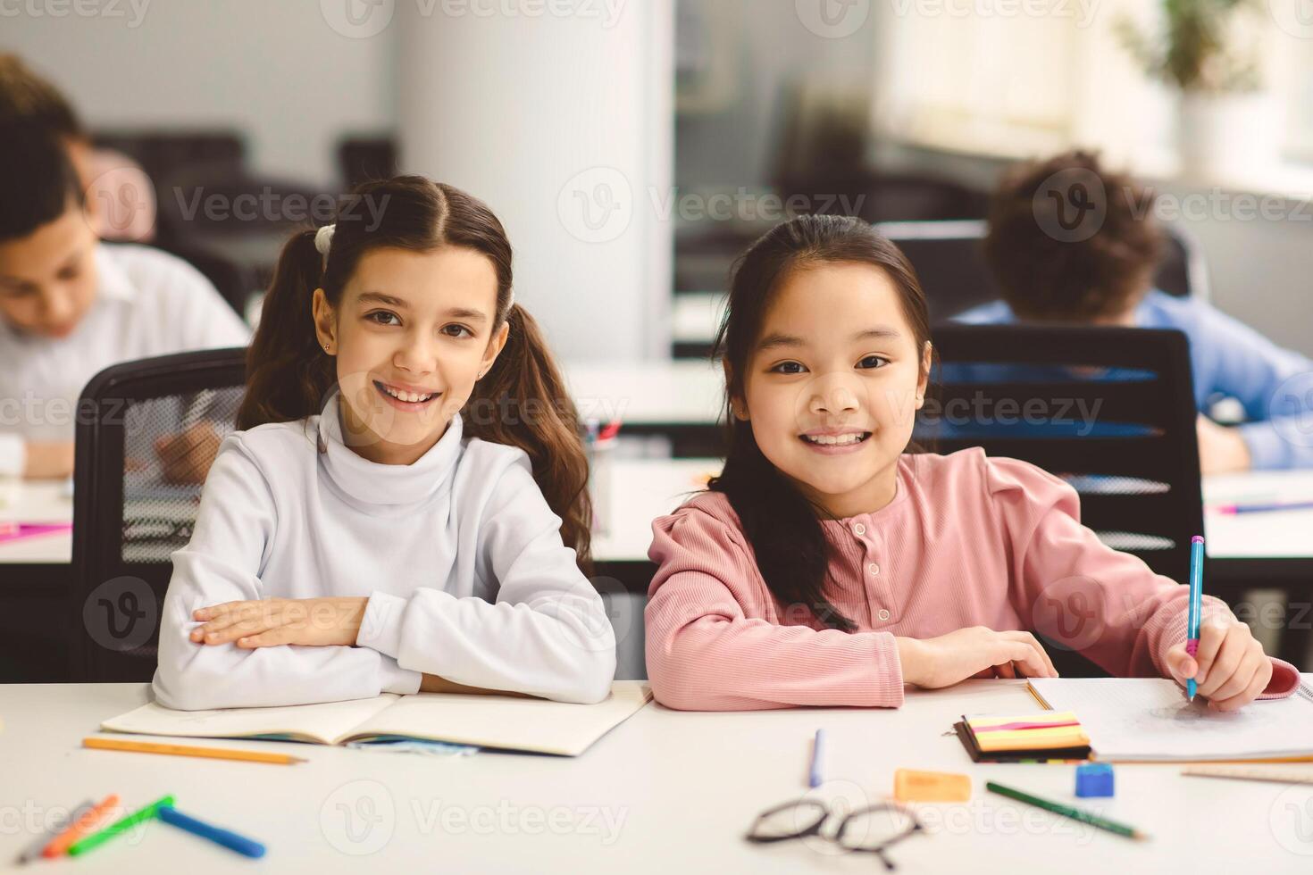 Education And Children Concept. Portrait of happy smiling small girls sitting at desk in classroom at elementary school with group of classmates in background and posing, looking at camera photo