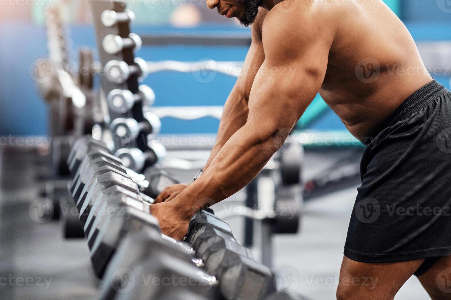 Shirtless african american muscular bodybuilder grabbing dumbbell at modern gym, copy space. Handsome black athletic man working out with dumbbells, making work out with weights photo