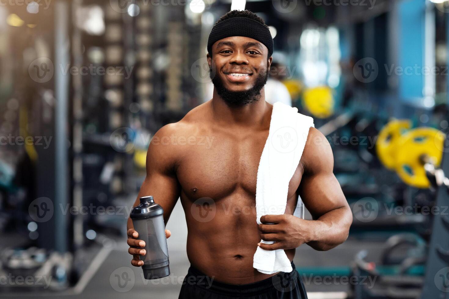 Rest after workout. Happy african american bodybuilder holding water or protein and towel at gym, empty space. Smiling black muscular man having break while exercising at modern gym photo