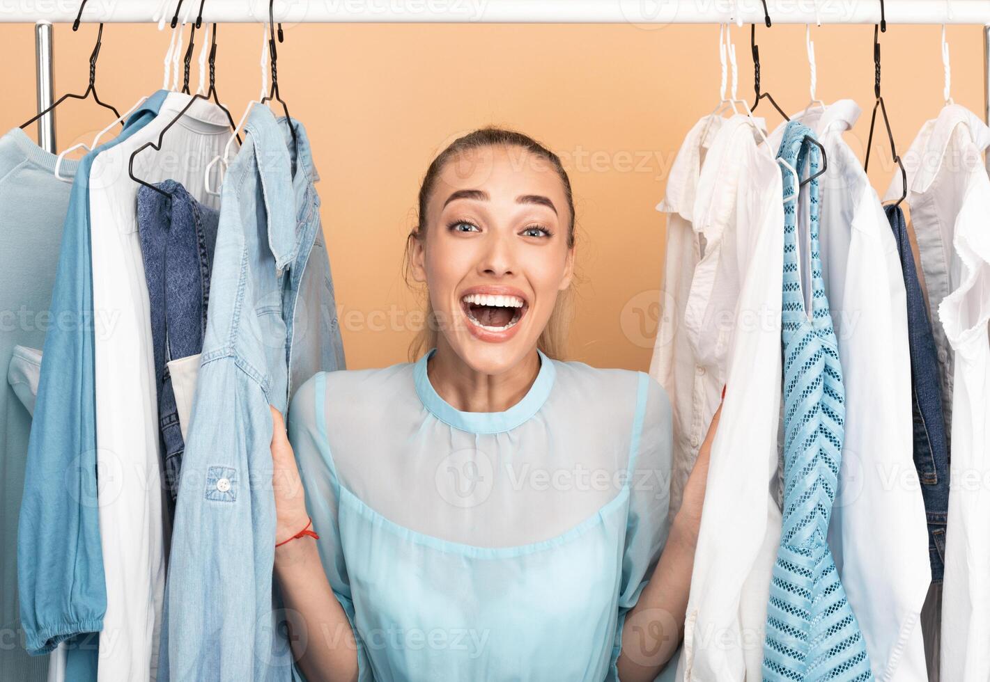 Shopaholic Concept. Portrait Of Emotional Young Woman Standing At Clothing Rail In Closet. Female Customer Shouting And Screaming With Joy, Choosing Clothes Over Pastel Studio Background photo