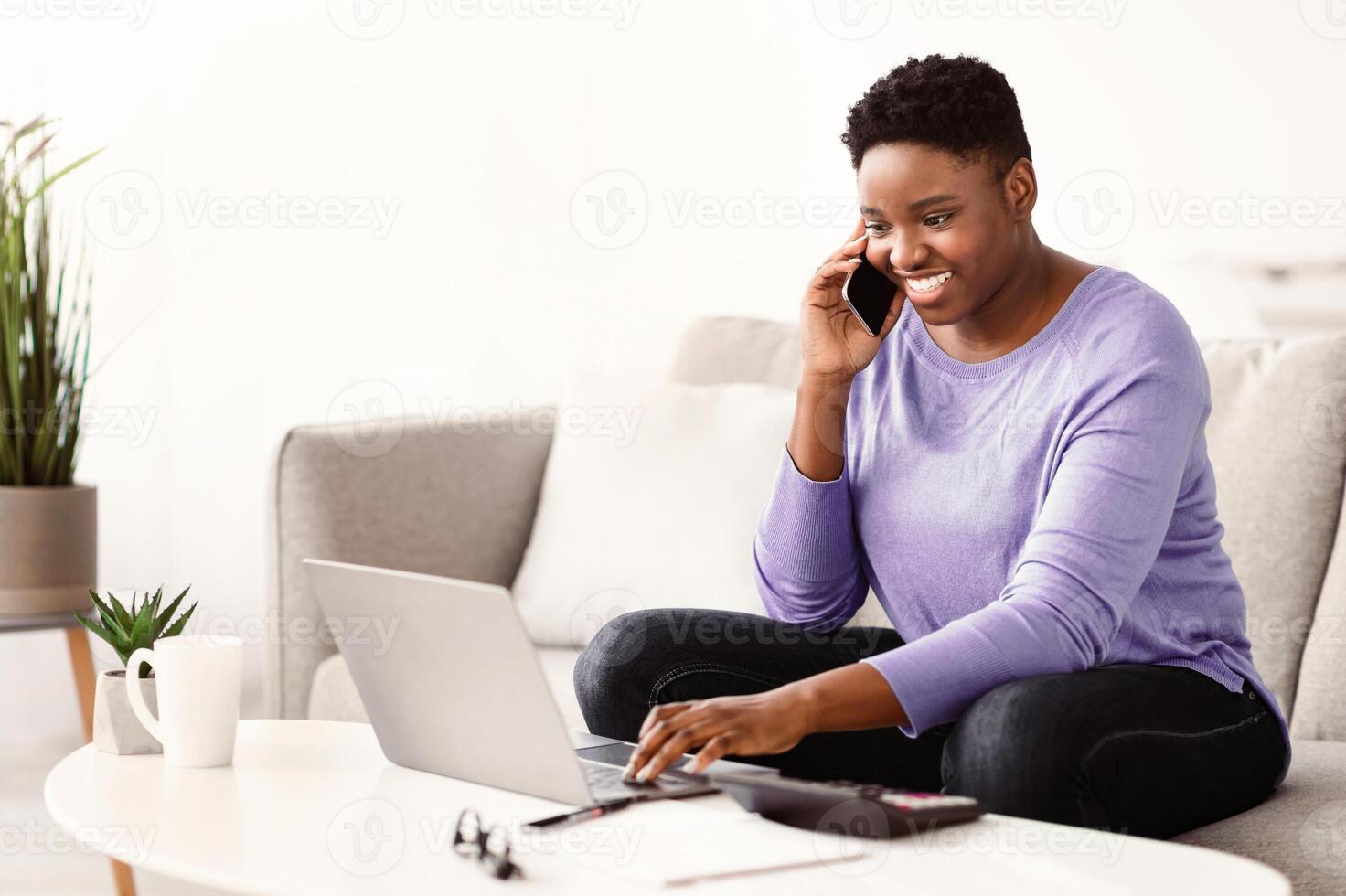 Multitasking At Home. Smiling young plus size black lady making call using her smartphone, sitting on sofa, typing on laptop keyboard. Positive woman talking with client or colleagues about work photo