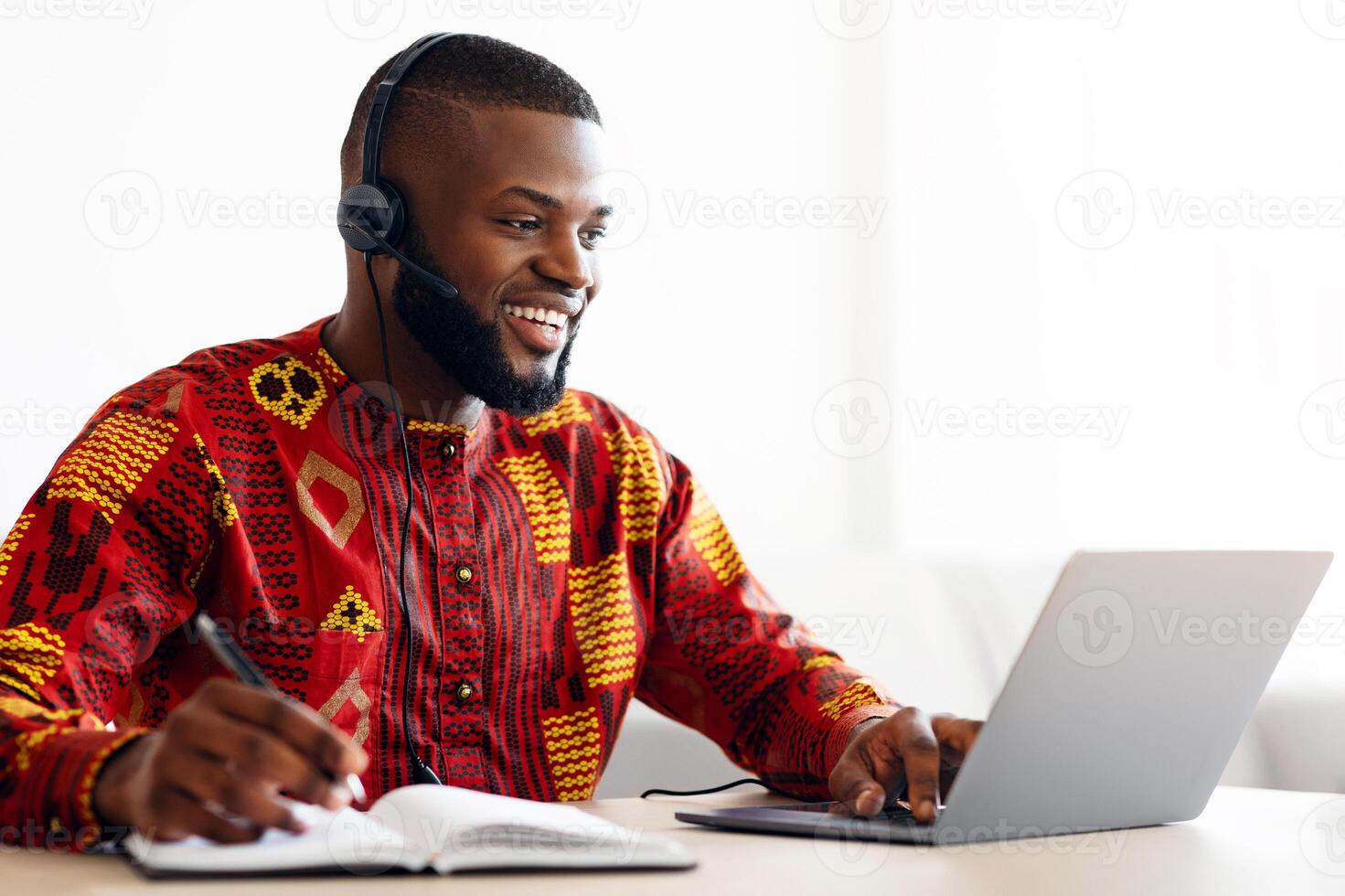 Online Education. Young African Man In Headset And Traditional Clothes Study With Laptop, Watching Webinar On Computer And Taking Notes, Sitting At Desk At Home Having Internet Training , Free Space photo