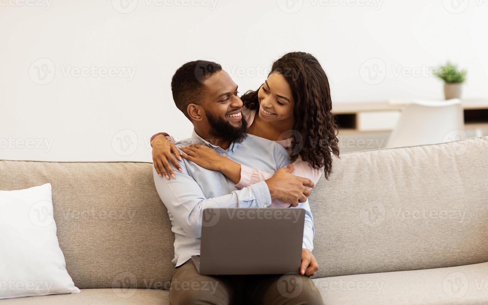 Family Happiness. Cheerful black couple web surfing on laptop together, sitting on sofa at home, relaxing and hugging, copy space. Smiling young african american woman embracing man from behind photo