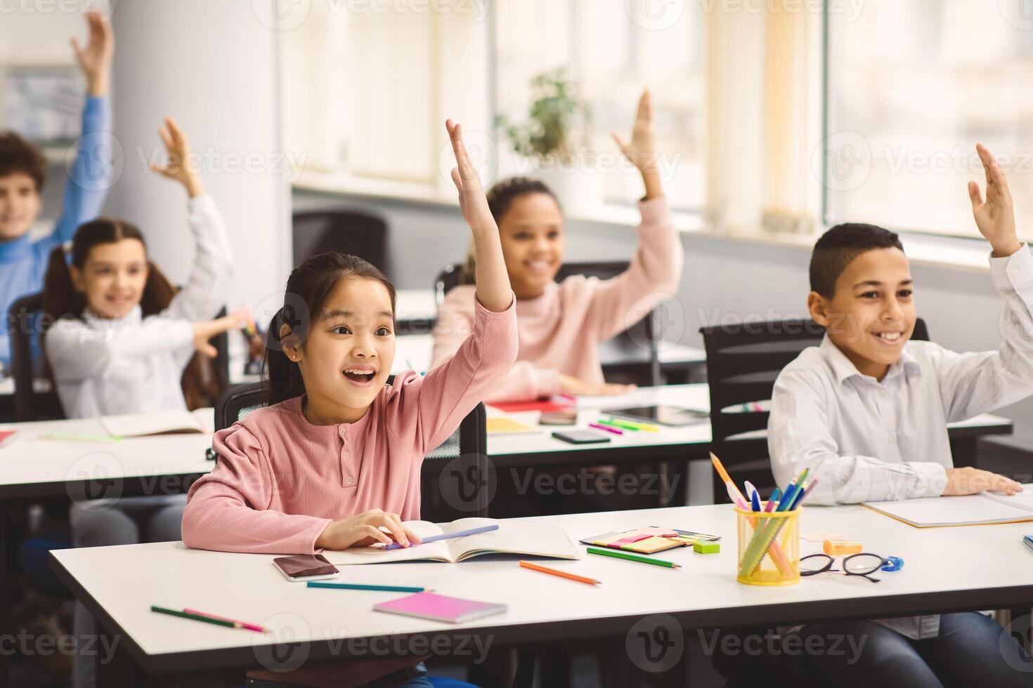Academic Concept. Diverse group of multiethnic smart junior schoolchildren sitting at desks in classroom and raising hands for an answer, studying with pleasure. Positive young elementary students photo