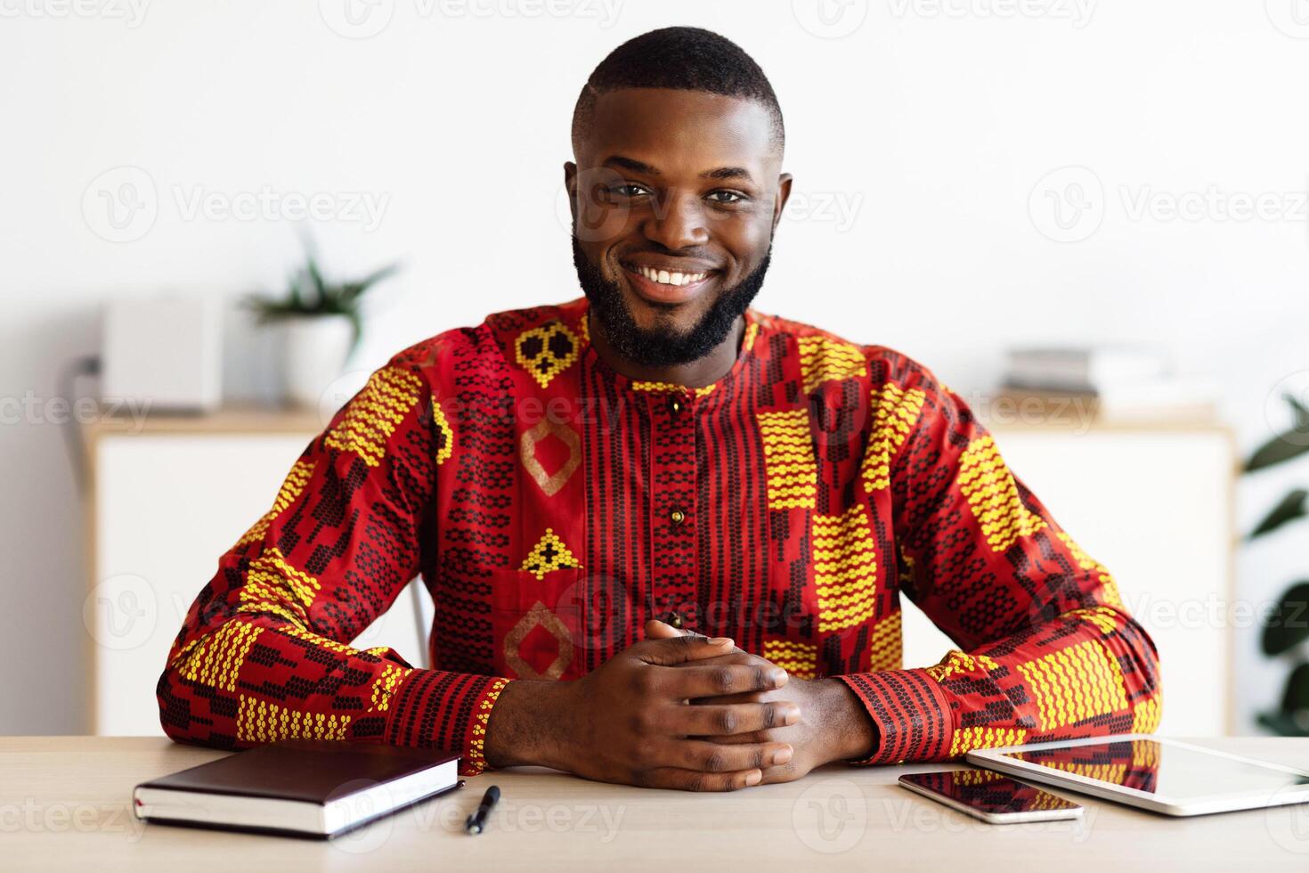 Portrait Of Handsome African Freelancer Man In Traditional Costume Sitting At Desk In Home Office, Positive Black Self-Employed Guy In Ethnic Costume Smiling And Looking At Camera, Free Space photo