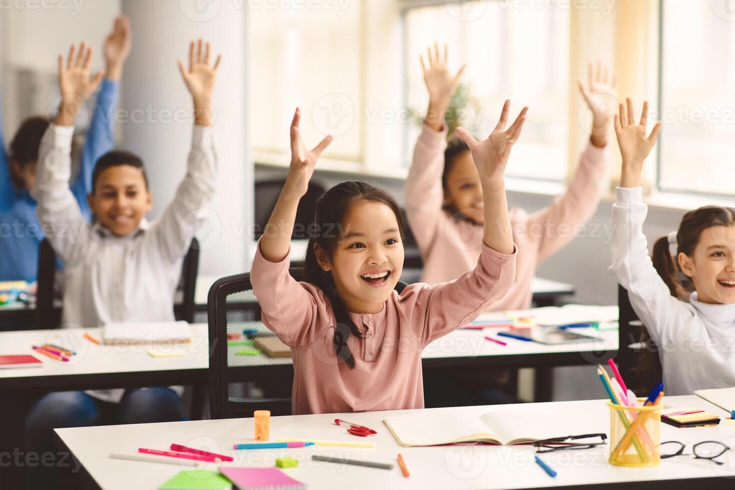 Portrait of multiciltural excited diverse group of small happy elementary school children sitting at desks in classroom, raising hands high up and waving, having fun, learning with pleasure photo