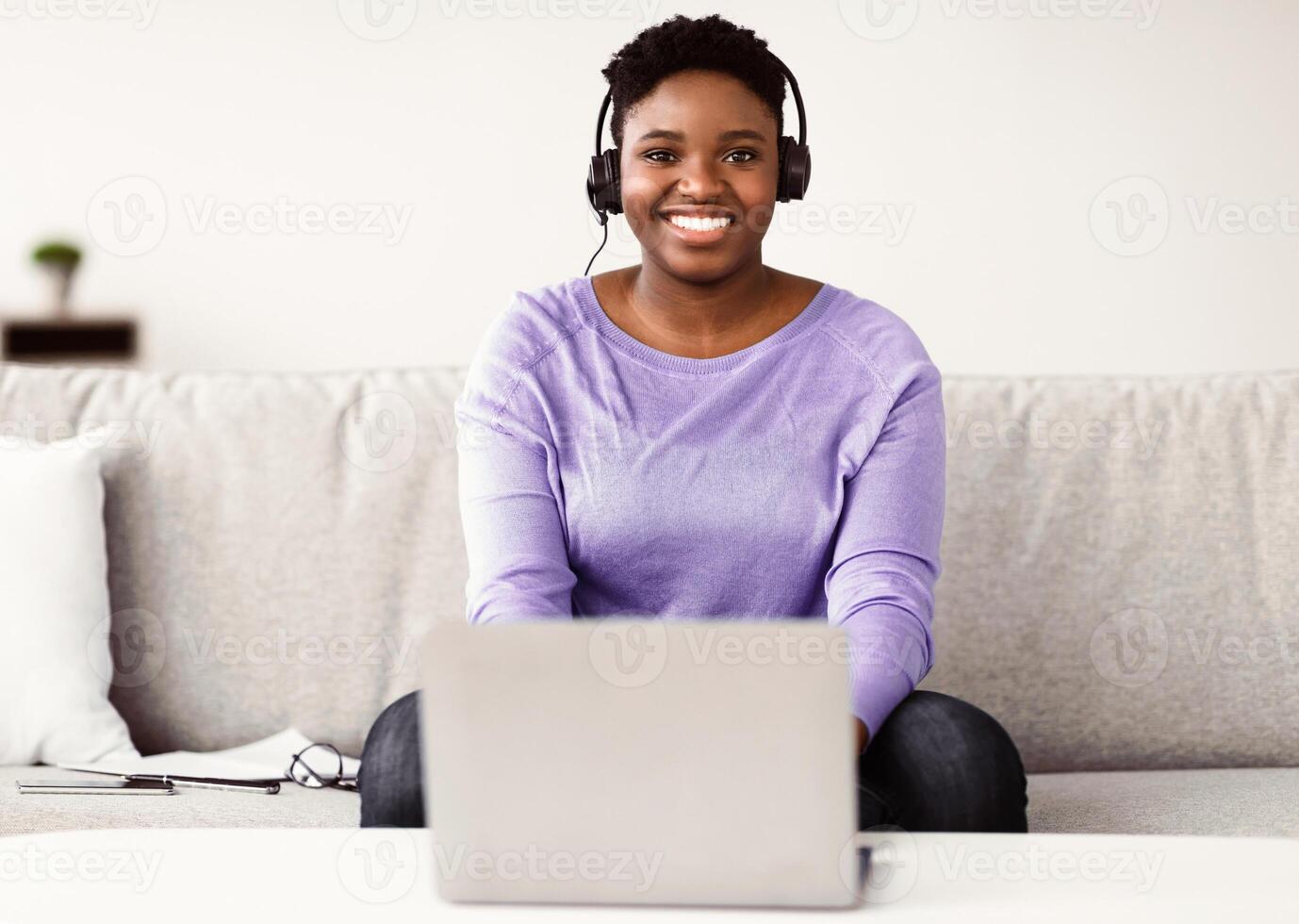 E-learning Concept. Happy black woman wearing headset studying online with laptop at home, cheerful casual african american lady sitting on sofa at desk with computer, looking at camera photo