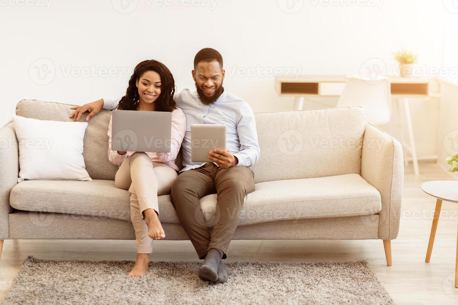 Family Gadgets. African American Couple Resting On Couch With Their Devices, Woman Using Laptop, Man Holding Digital Tablet, Browsing Internet Together At Home. Modern Lifestyle Concept, Copy Space photo