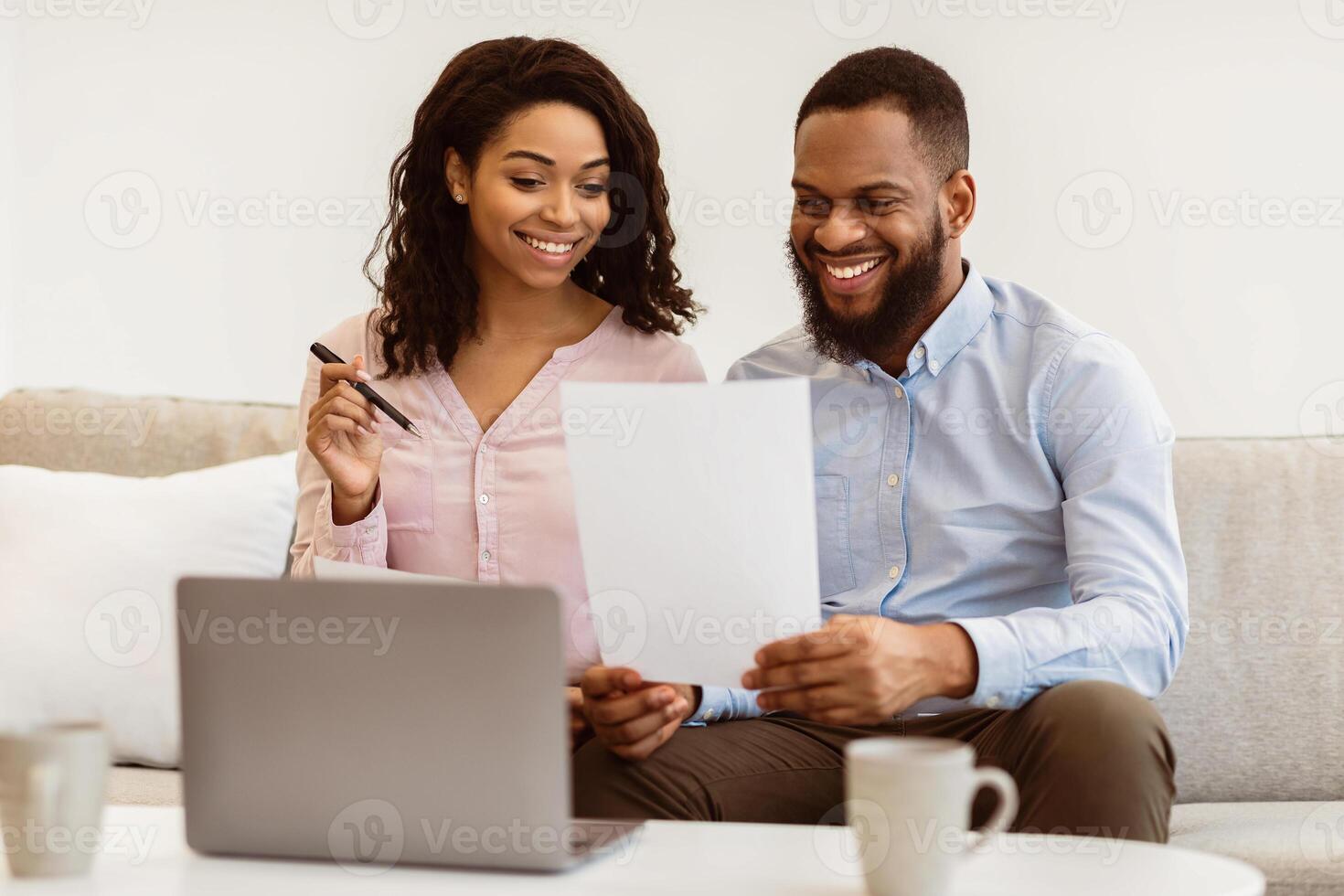 Family Business And Accounting. Cheerful black couple holding paper, checking household expenses, reading documentation or receipt from bank, sorting bills or planning budget, sitting on couch photo