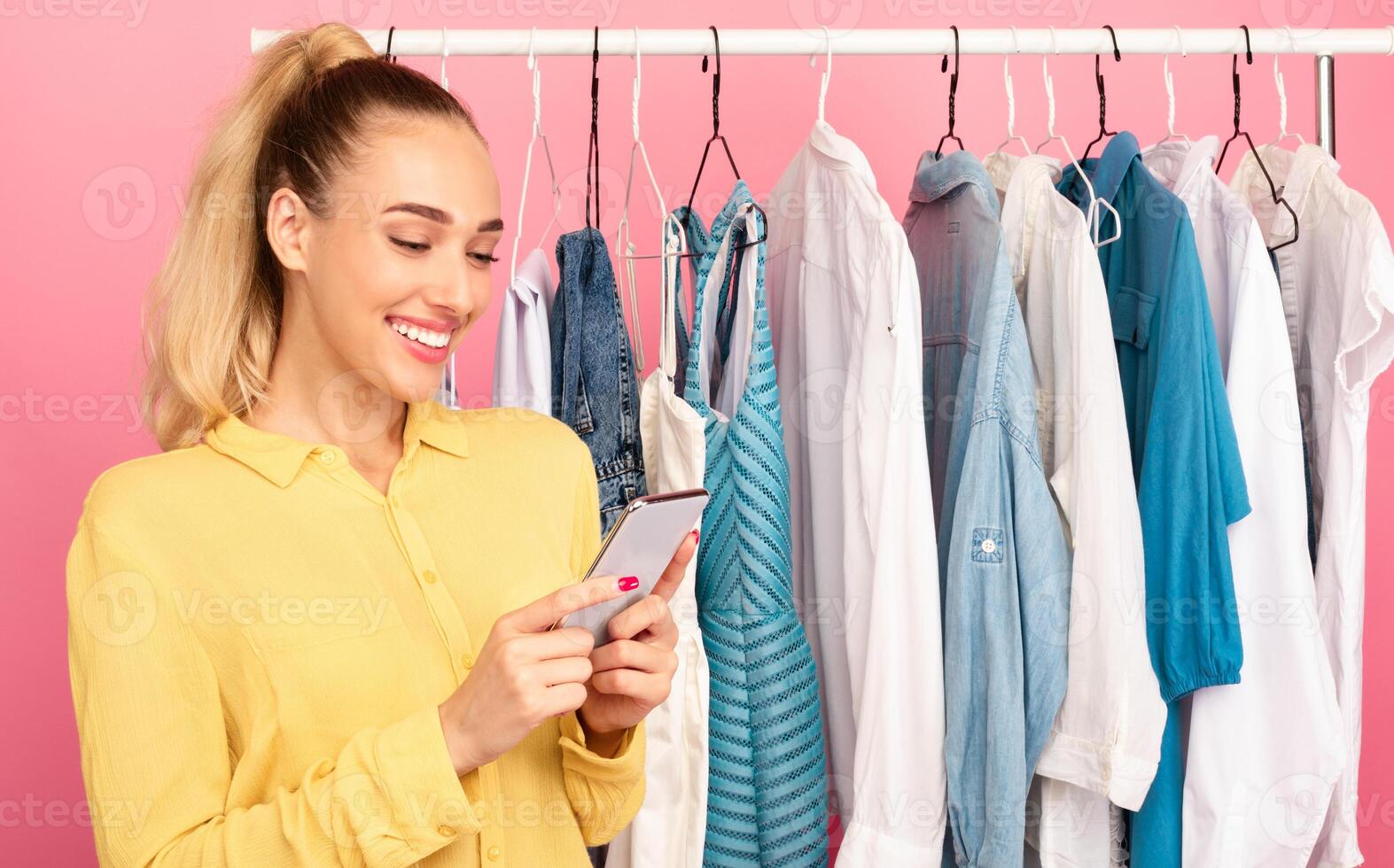 Portrait of smiling fashion blogger influencer holding and using her smartphone, texting sms, checking social media, writing post or making purchases in online store standing near clothing rail photo
