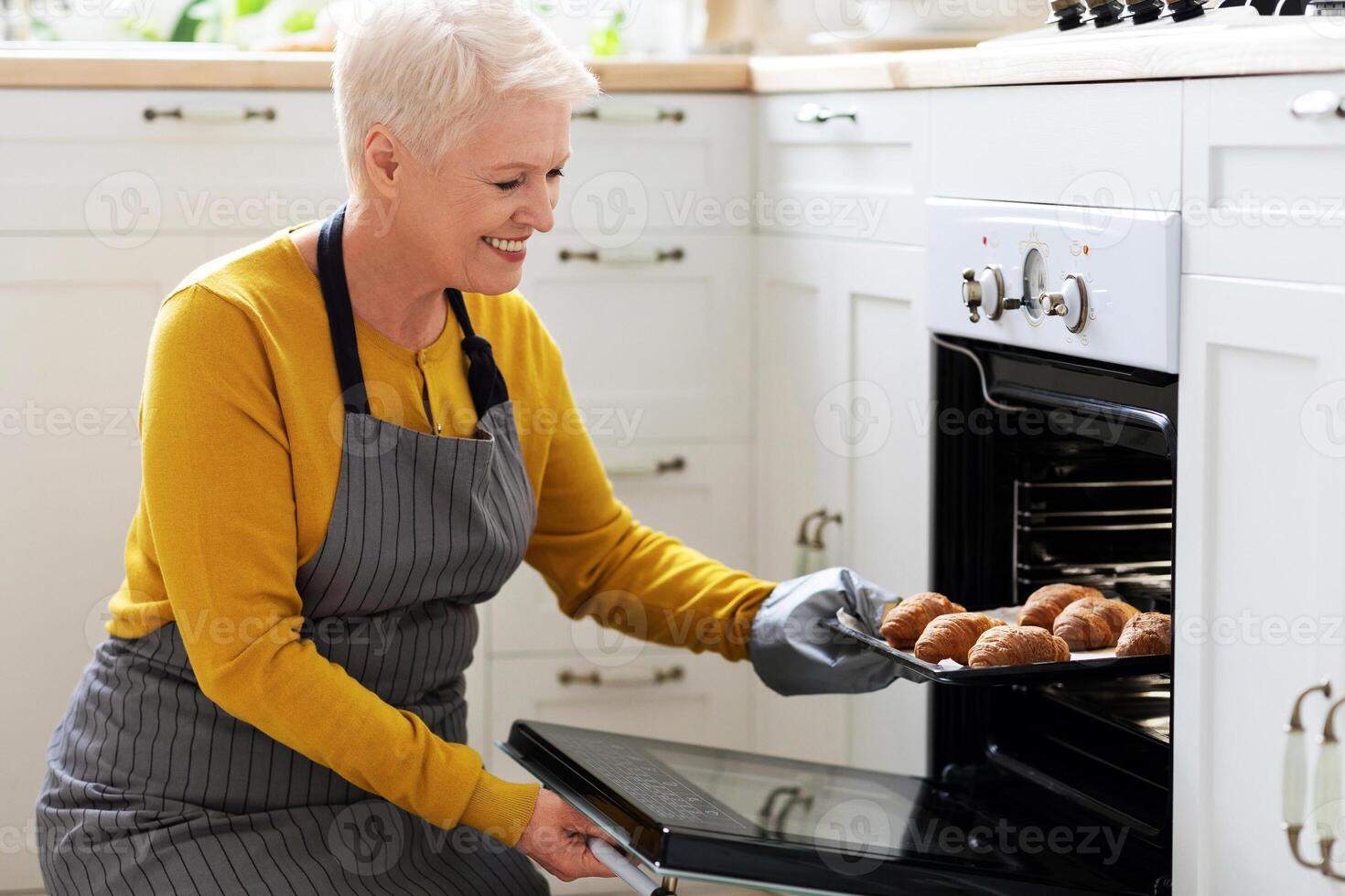 Smiling elderly woman baking pastry at home, taking delicious croissants out of oven, copy space. Joyful senior lady in apron opening oven, checking her pastry, cozy kitchen interior, closeup photo