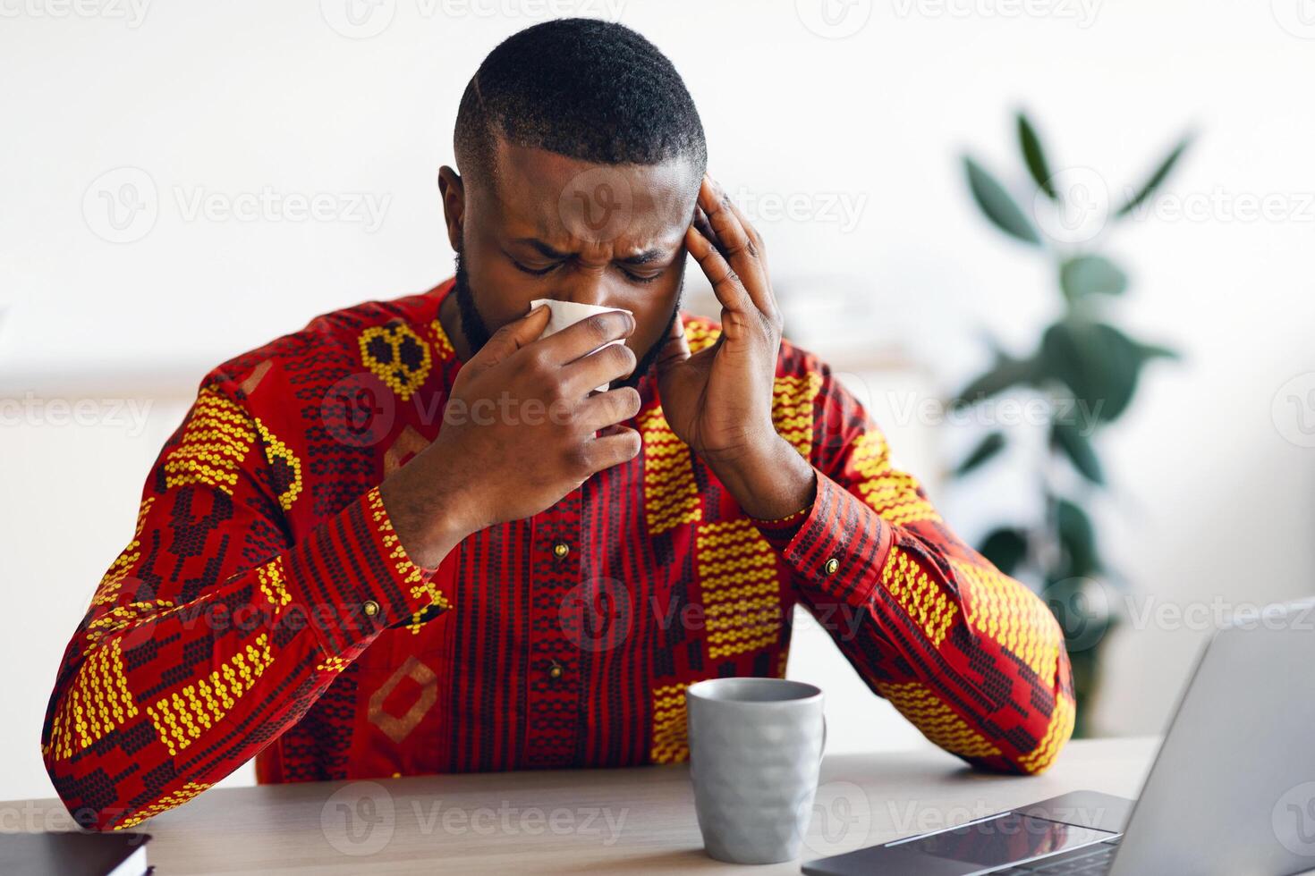 Sick African Man In Traditional Shirt Feeling Unwell While Working In Office, Sitting Desk With Laptop And Blowing Nose In Paper Napkin. Ill Black Guy Suffering Coronavirus Symptoms, Free Space photo
