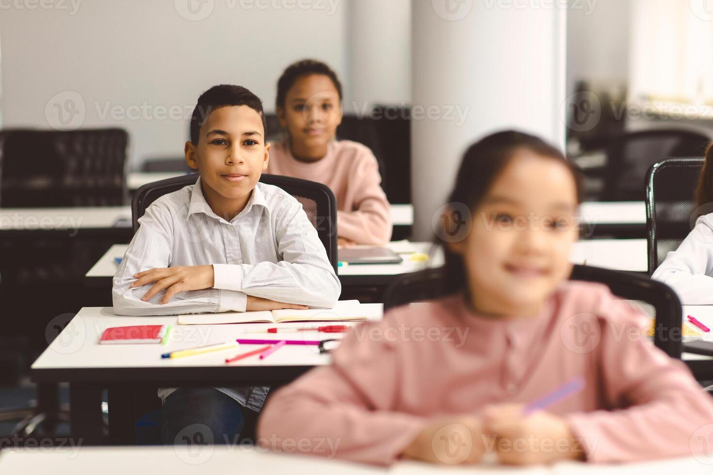 Elementary Education. Portrait of small smiling boy sitting at table in classroom at primary school or kindergarten with group of classmates. Return back to school after covid quarantine photo