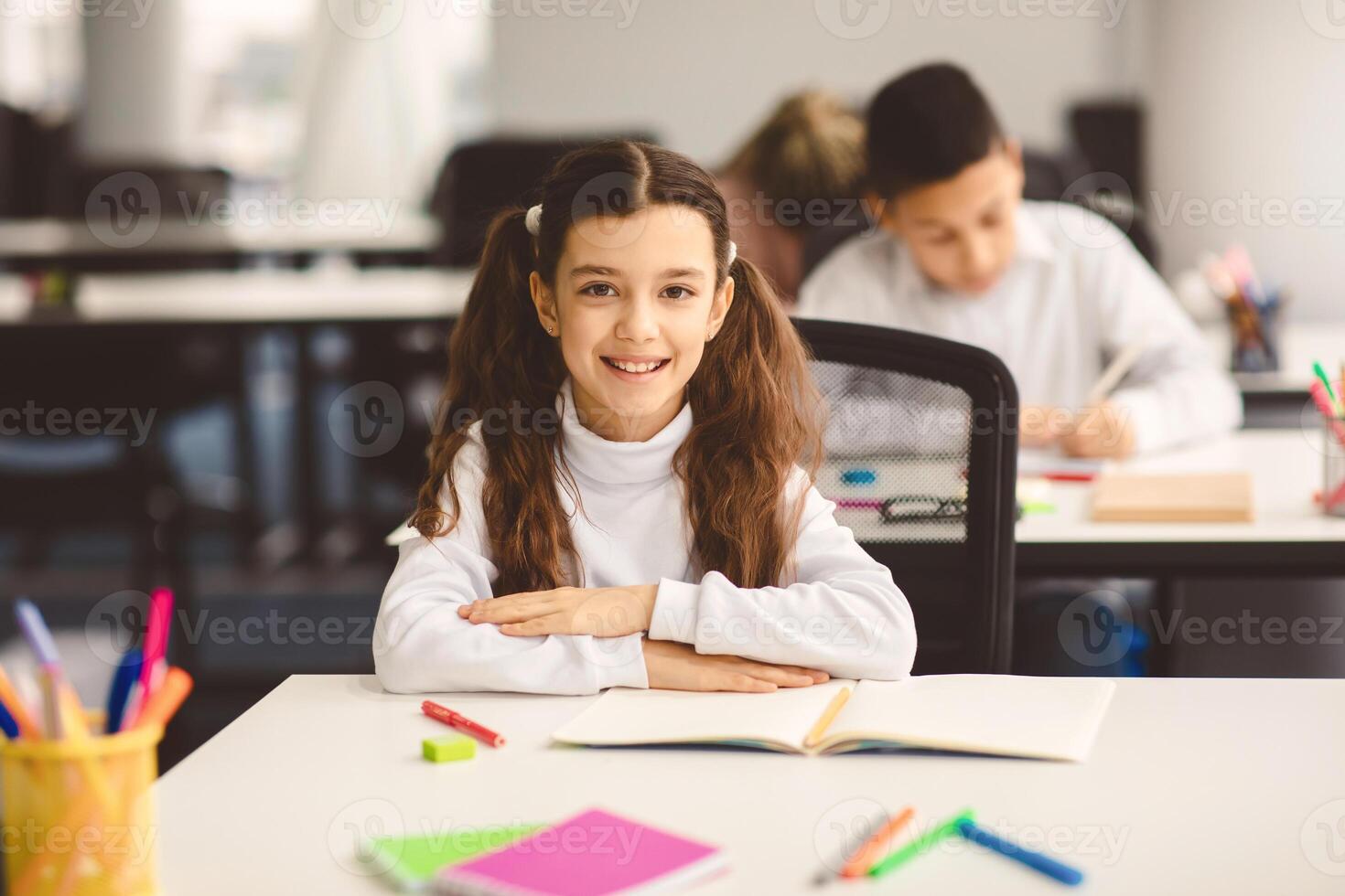 First Day At School. Portrait of smiling diligent small girl with ponytails sitting at table in classroom at elementary school with group of classmates in background and posing, looking at camera photo