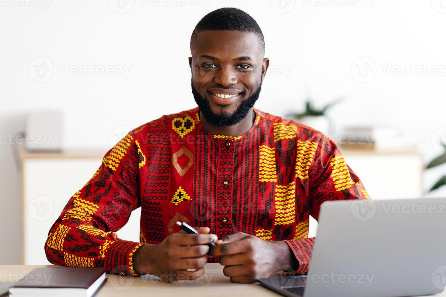 Handsome African Freelancer Guy In Traditional Costume Sitting At Desk At Home Office, Working On Laptop Computer And Smiling At Camera, Black Man Enjoying Remote Work And Freelance Career, Free Space photo