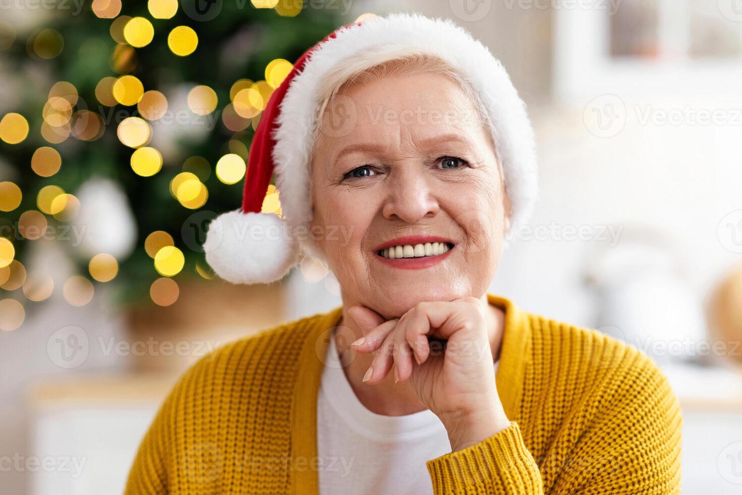 Portrait of cheerful old woman in Santa hat smiling at camera, celebrating Christmas at home, closeup. Happy senior lady sitting at kitchen decorated with xmas tree, smiling at camera photo