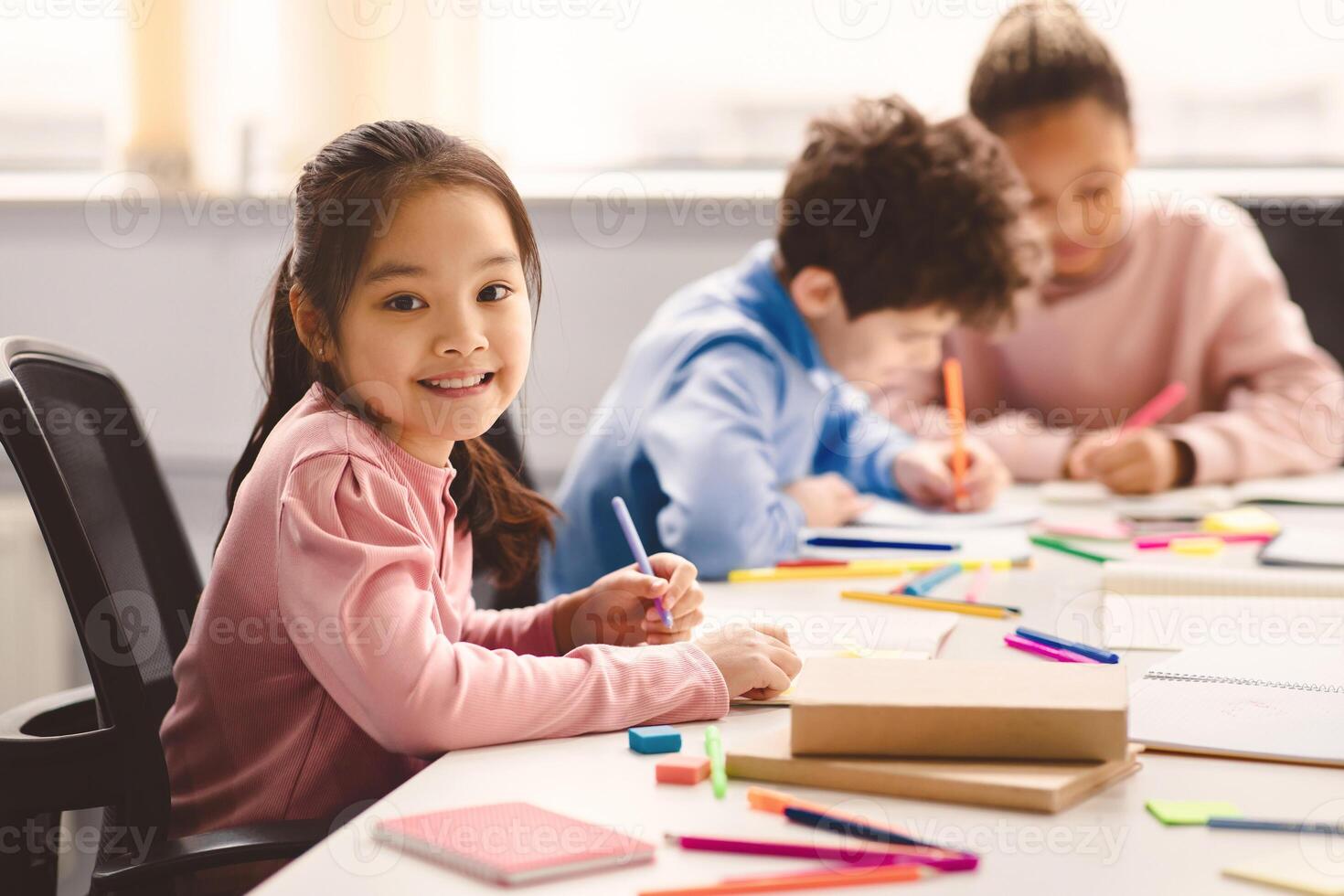 Academic Concept. Smiling junior asian school girl sitting at desk in classroom, writing in notebook, posing and looking at camera. Group of diverse classmates studying in the background photo