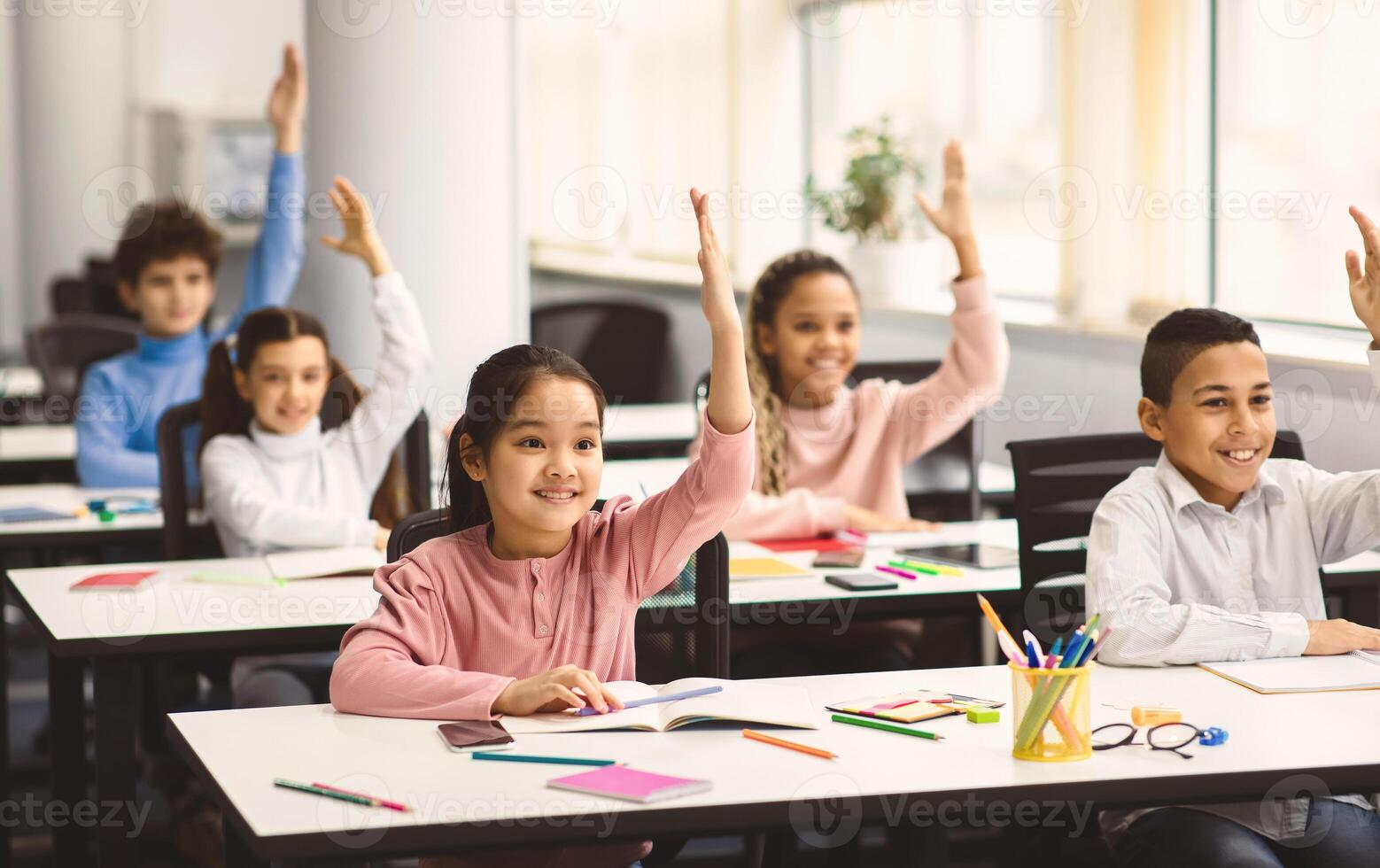 Return Back To School. Multiciltural diverse group of happy small elementary schoolchildren sitting at desks in classroom, raising hands up for an answer, learning with pleasure. Social distance photo