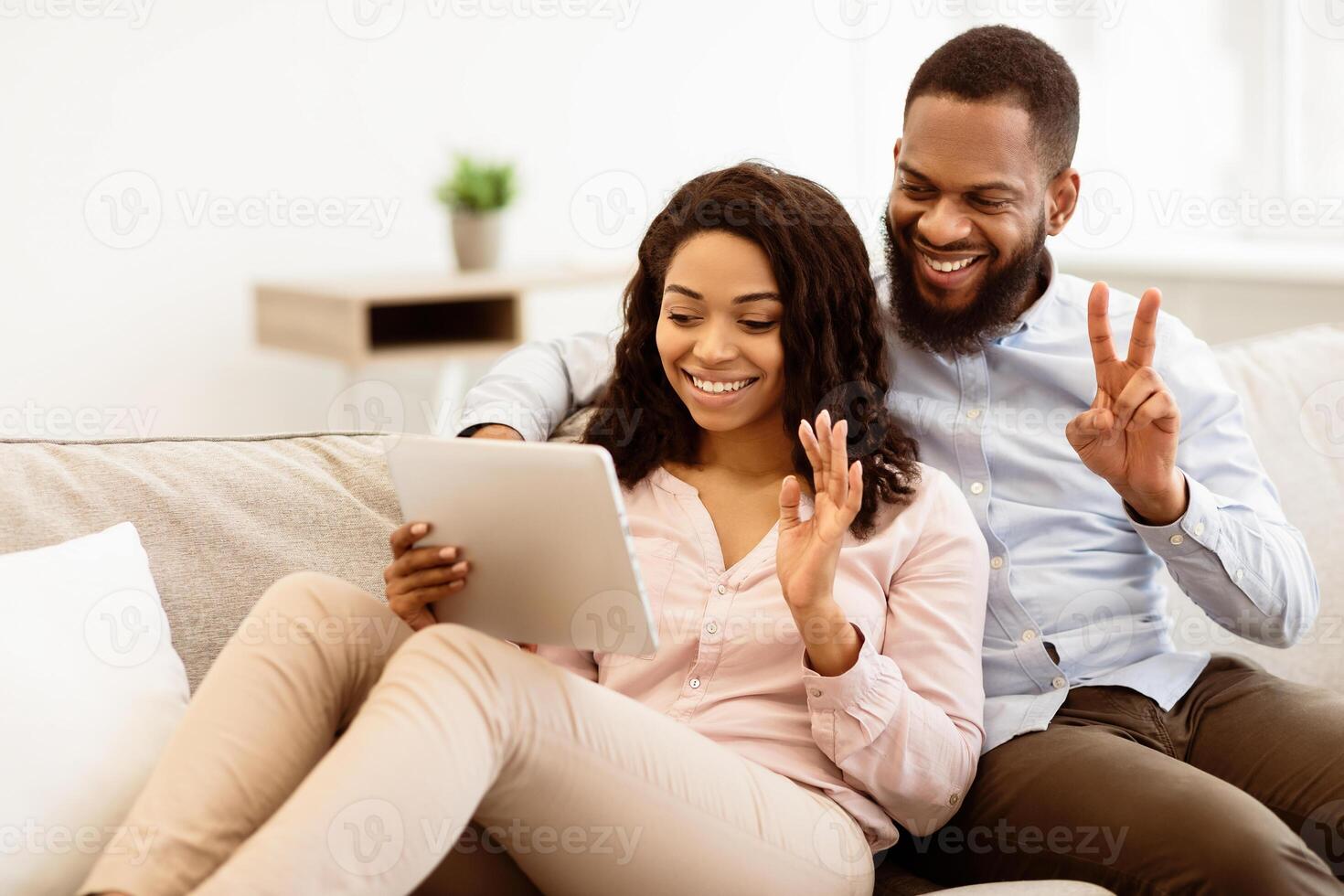 Online Communication. Smiling african american man and woman making digital chat with friends or family using tablet, waving to webcam, sitting and resting on the couch in living room photo