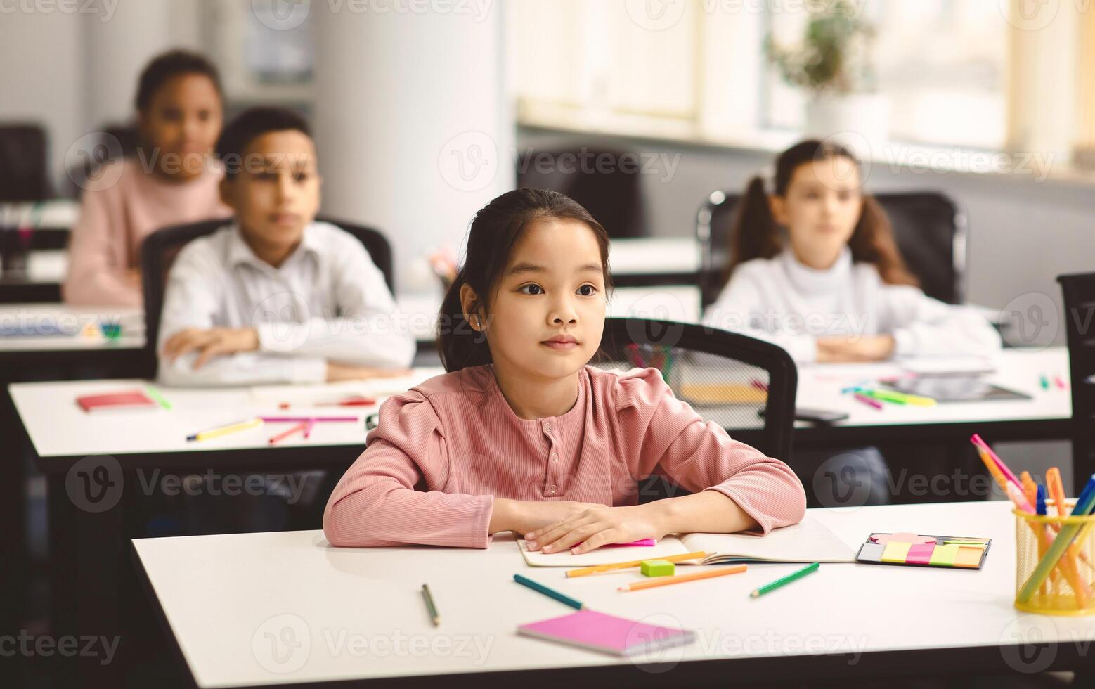 Return Back To School. Portrait of focused diligent asian female pupil sitting with folded arms at desk with group of diverse classmates, listening to teacher, selective focus. Academic Concept photo