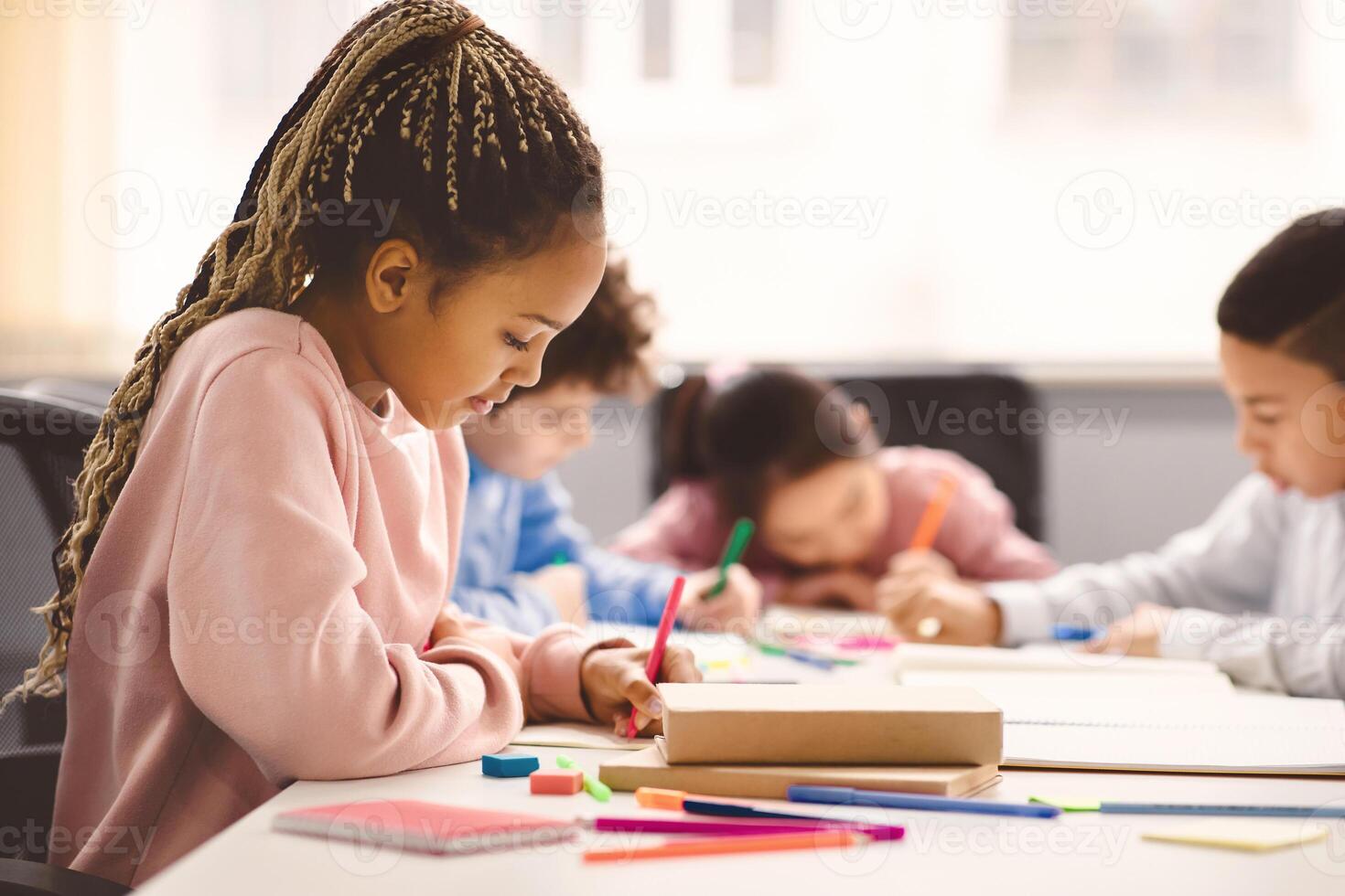 Portrait of small black girl with afro braids sitting at table in classroom at primary school or kindergarten, writing or drawing in notebook. Reopening and return back to school after lockdown photo