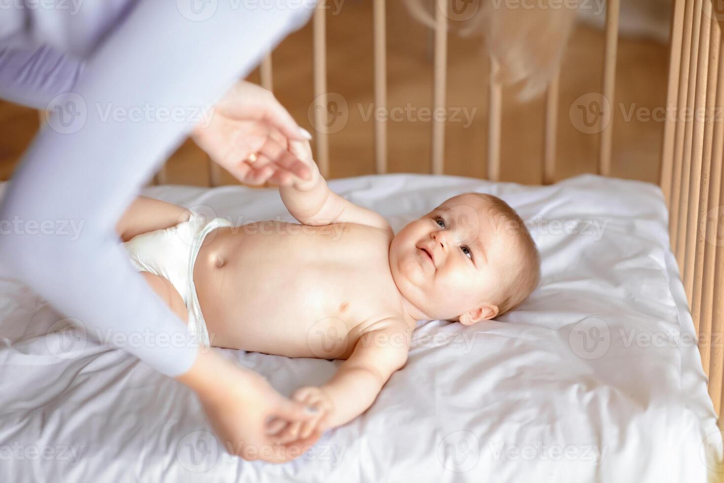 Good morning sunshine. Smiling little baby laying in cot, holding mom's hands, ready to wake up. Top view of cute blonde toddler waking up, lying in wooden baby bed, grabbing mother fingers photo