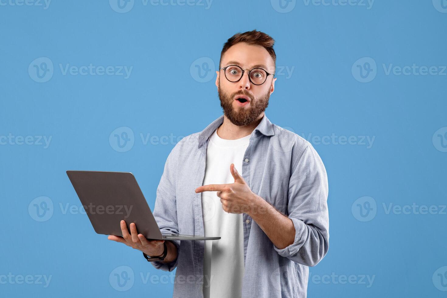 Surprised millennial guy opening mouth in awe, pointing at laptop screen with shocking web content on blue studio background. Young man watching unbelievable breaking news photo