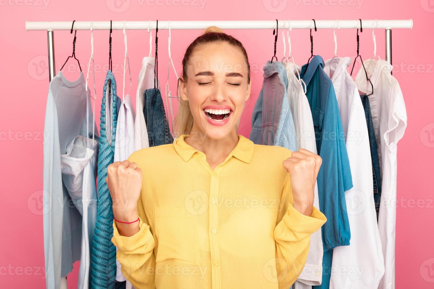 Sales And Discounts. Portrait Of Emotional Young Lady Screaming Yes With Joy, Closing Eyes, Shaking Clenched Fists. Female Customer Laughing And Gesturing, Clothing Rail In The Background photo