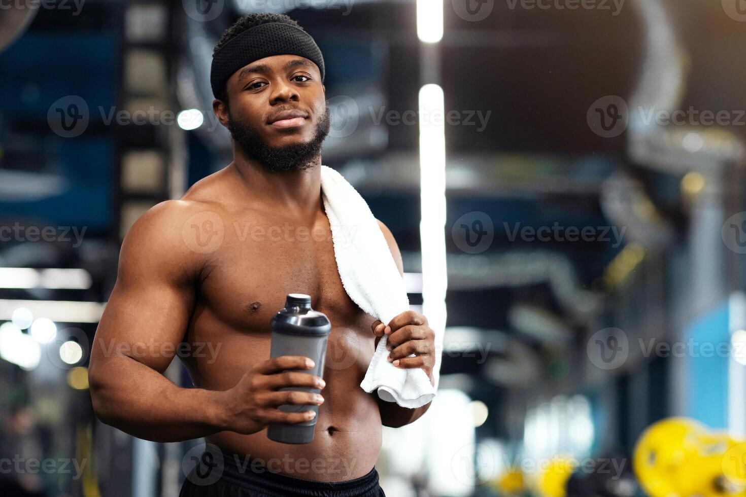 Rest after workout. Shirtless african american bodybuilder holding water or protein drink and towel at gym, empty space. Handsome black muscular man having break while exercising at modern gym photo