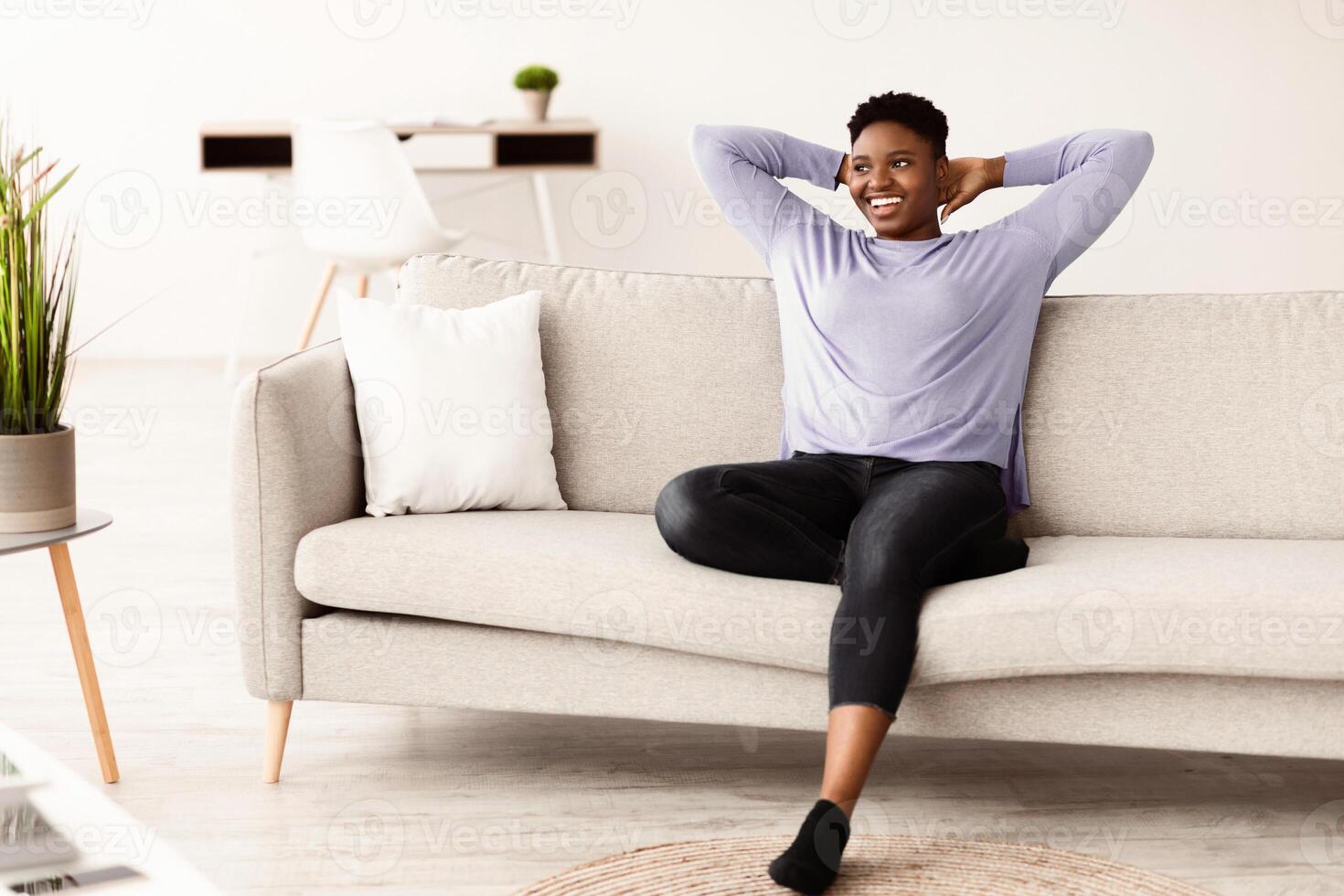 Smiling curvy young black woman with hands behind head relaxing on cozy sofa at home, daydreaming and looking away, satisfied serene girl leaning back, stretching on comfortable couch in living room photo