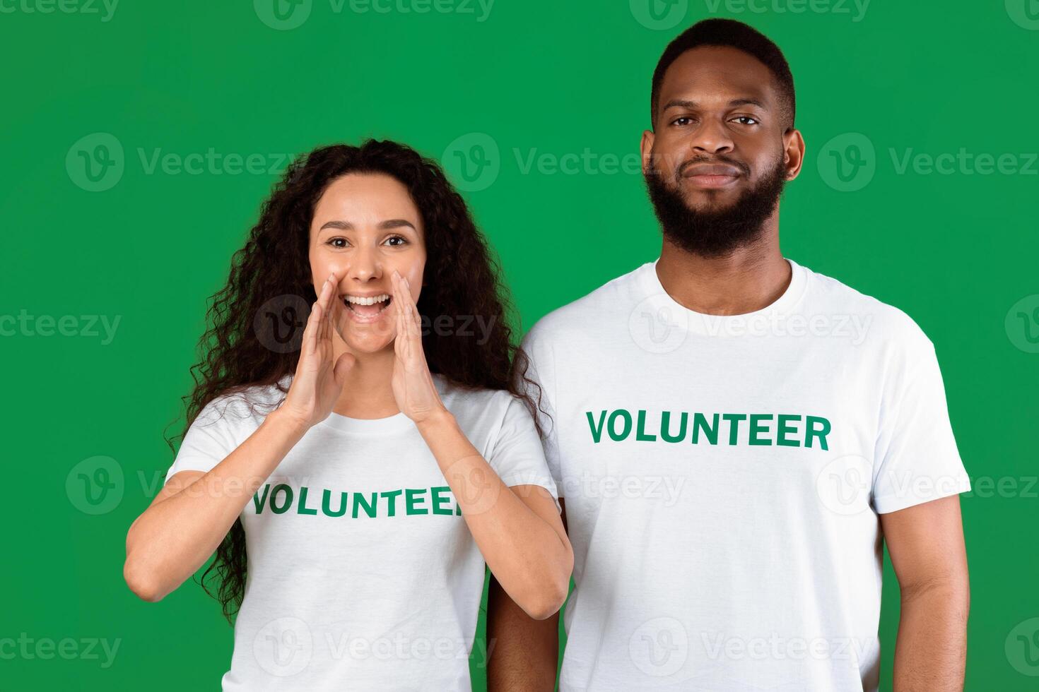 Two Multiethnic Volunteers Shouting Hey Holding Hands Near Mouth, Drawing Attention And Calling You Standing Posing Over Green Background. Volunteering Advertisement. Studio Shot photo