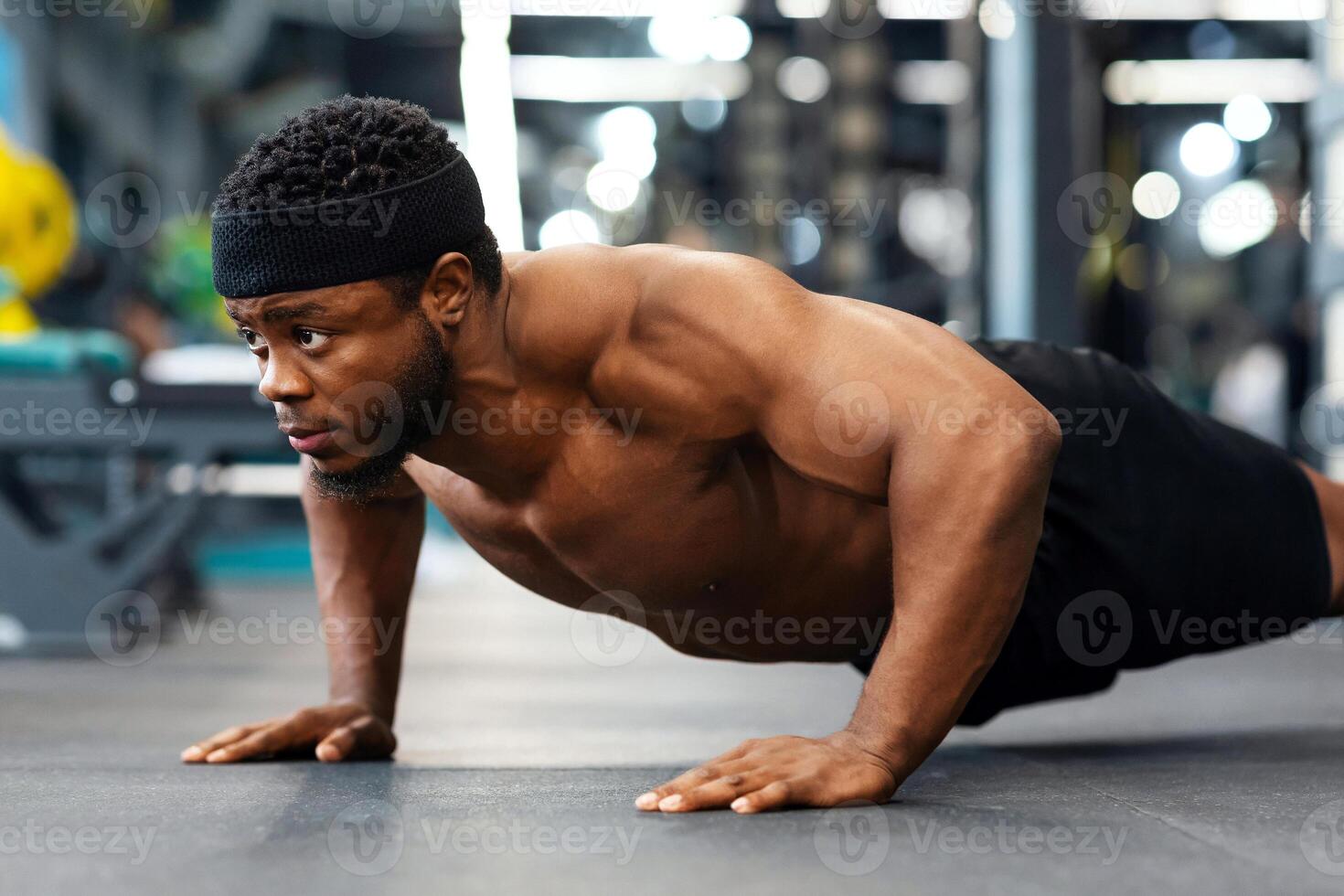 Muscular shirtless black man making push-ups on floor in modern male gym, side view. Motivated african american guy bodybuilder making work out for arms and chest on floor photo