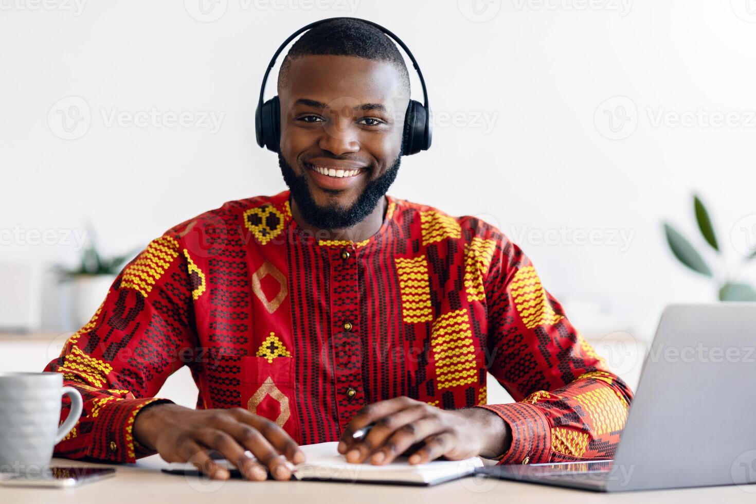 E-learning Concept. Happy black man wearing headset study online with laptop at home, cheerful african guy in traditional costume sitting at desk with computer and notepad, enjoying distance education photo