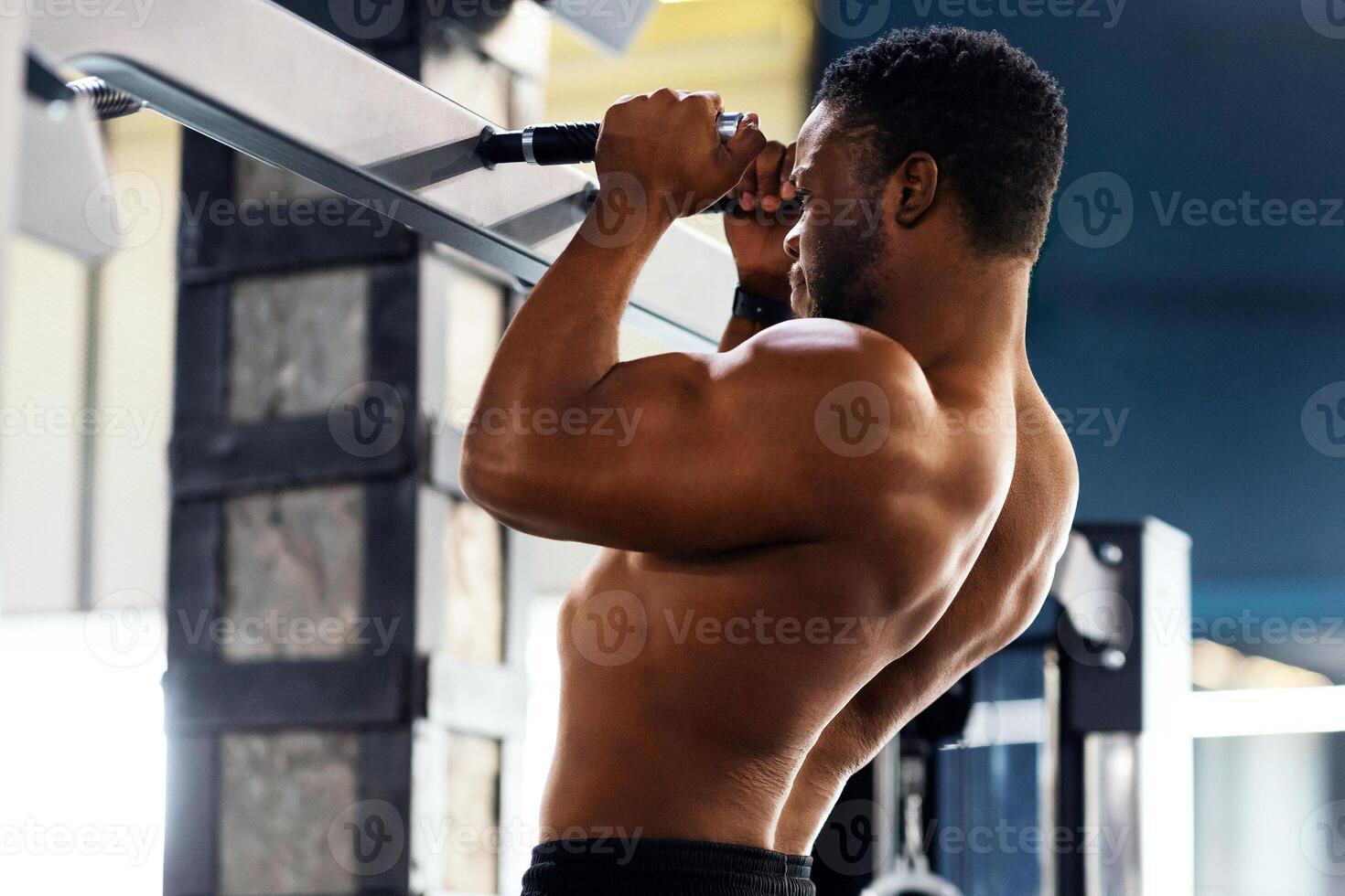 Muscular black guy athlete pulling up on horizontal bar at gym, free space. Shirtless african american sportsman bodybuilder doing pull-ups, working on arm muscles, side view photo